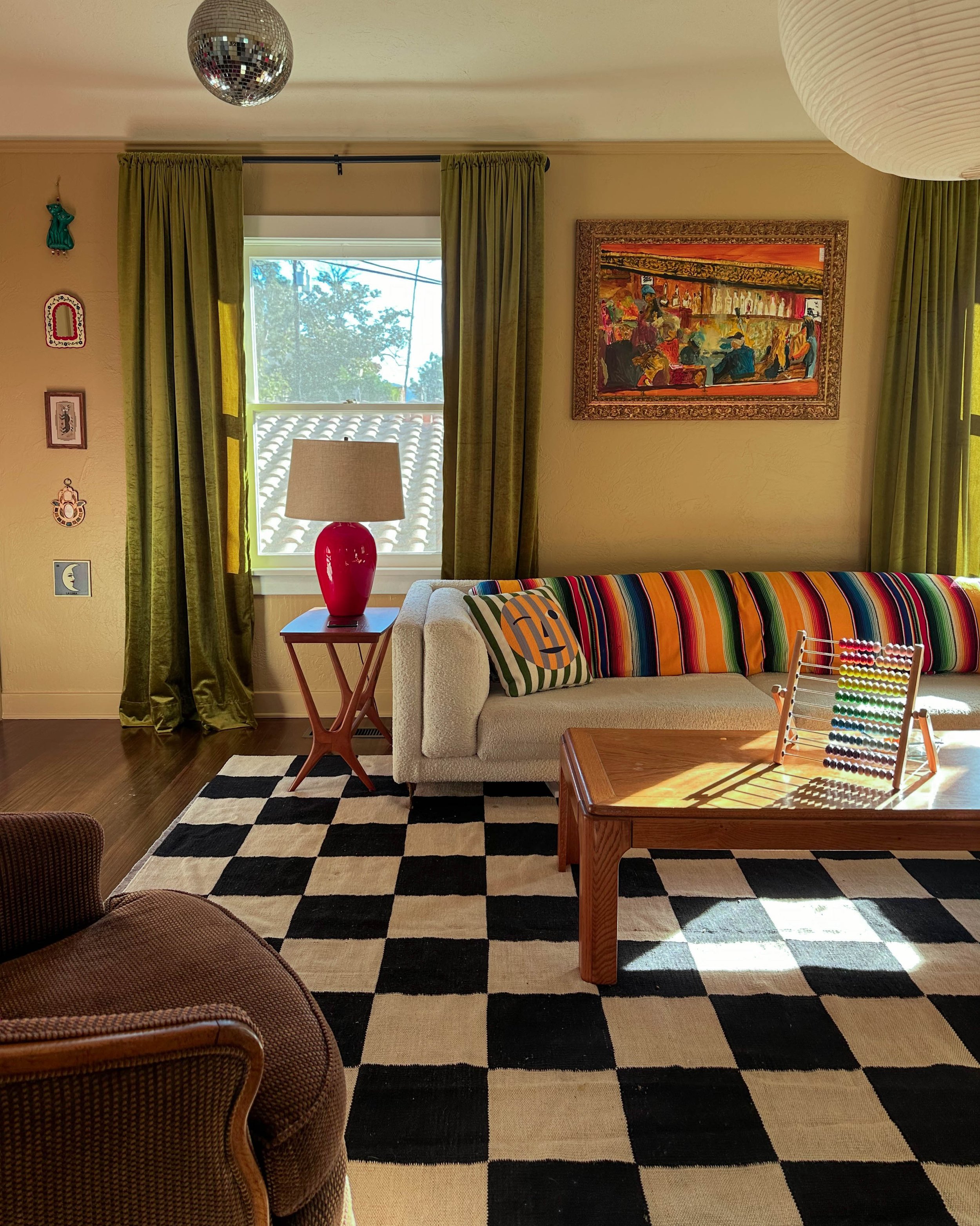 A cozy living room with a black and white checkered rug, beige couch with colorful striped pillows, a wooden coffee table with a rainbow abacus, a side table with a red lamp, and green curtains. There's a window showing blue sky and trees, framed artwork on the wall, and a small chandelier hanging from the ceiling.