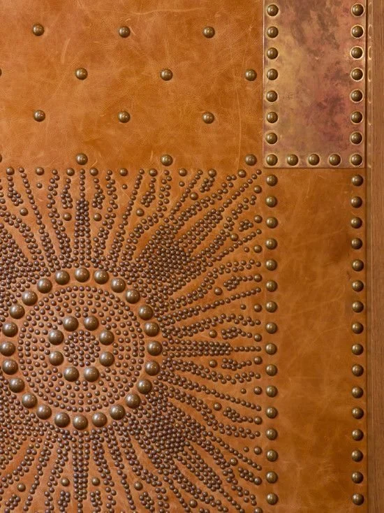 Close-up of a decorative metal surface with a spiral pattern of small round rivets or nails, surrounded by geometric arrangements of rivets on a brown, textured background.