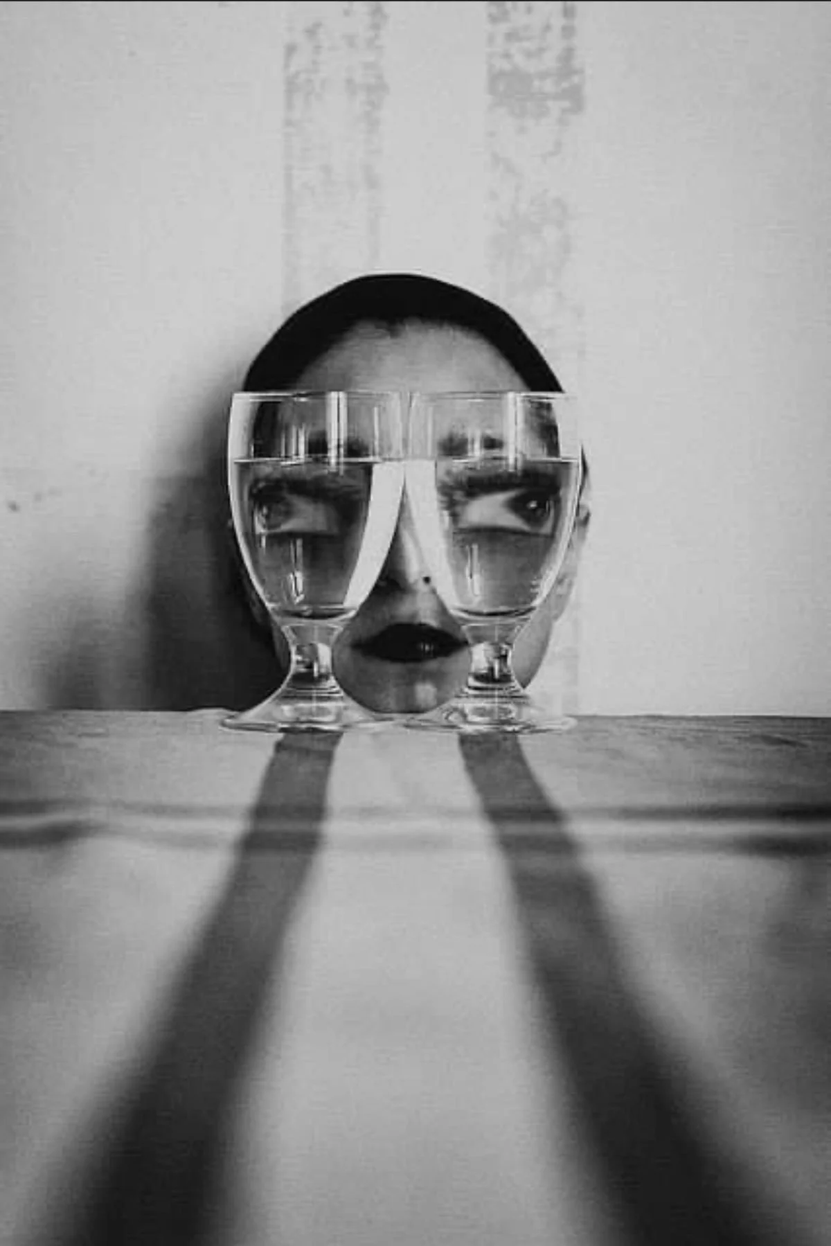 Schwarz-weiß-Foto eines Gesichts, das durch zwei Wassergläser auf einem Tisch zerlegt wird, wobei die Augen, Nase und Lippen sichtbar sind, jedoch auf unheimliche Weise durch die Gläser verfremdet werden. Der Hintergrund ist eine bemalte Wand.