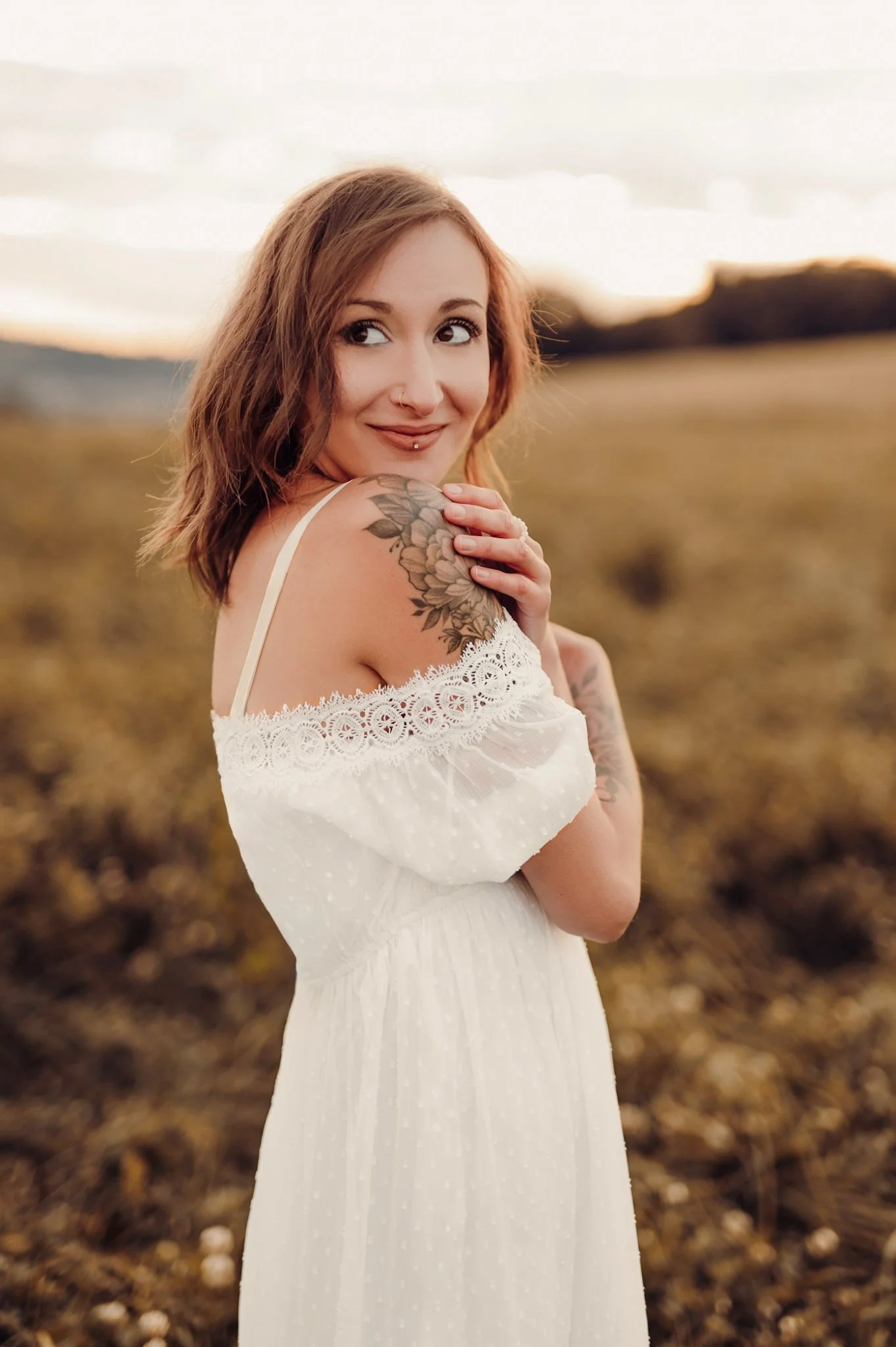Ein Portrait einer jungen Frau mit roten Haaren, Tattoos am Schulterbereich, in einem weißen Kleid, im Freien bei Sonnenuntergang in einer ländlichen Umgebung.