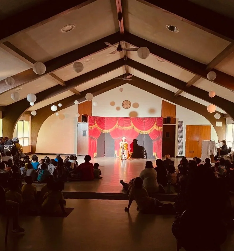 Rumplestiltskin performance by Wixie Children's Theatre in reception hall at RPCC. Photo is of an actor in costume sitting at a spinning wheel. In the shadowed foreground the audience is seated on the floor.