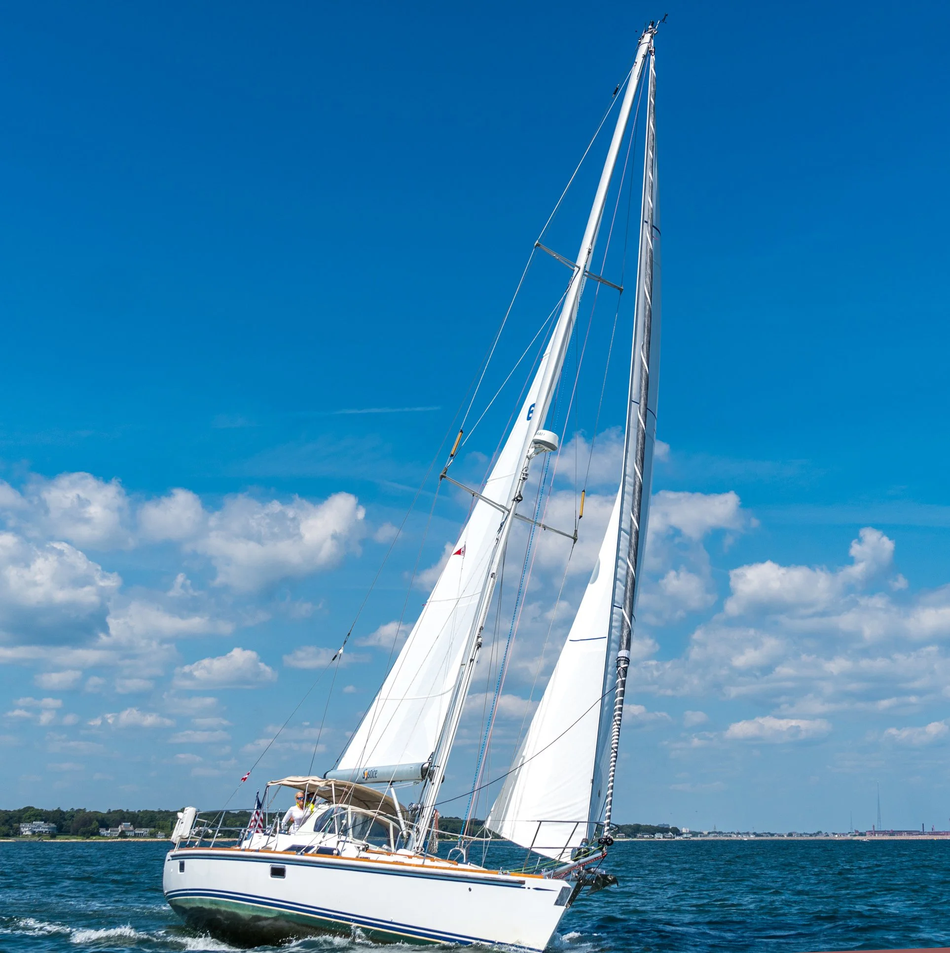 A white sailboat with its sails partially raised, sailing on calm blue water near a coastal town with houses and trees, under a clear blue sky with some clouds.