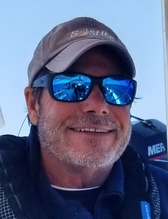 Smiling man outdoors wearing sunglasses, a cap, and a dark jacket.
