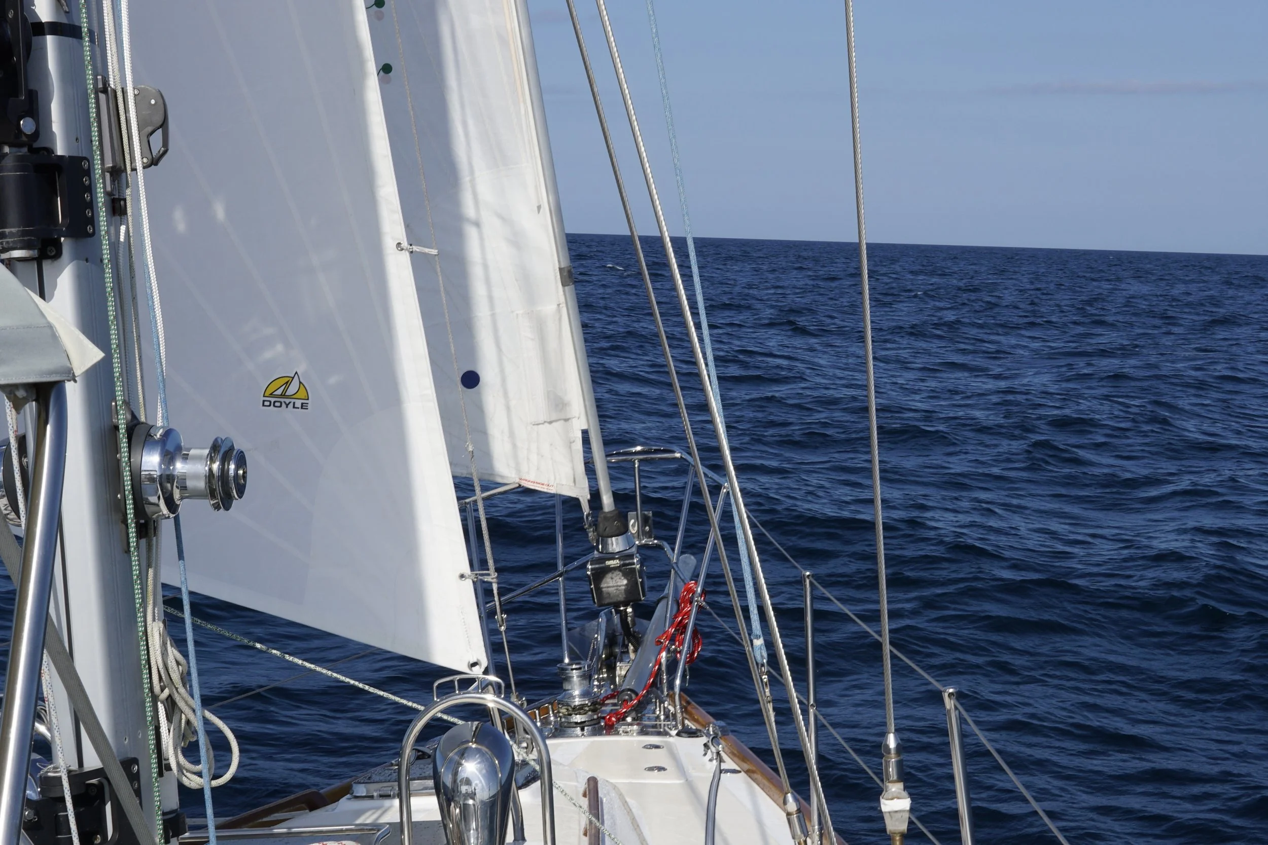 View from a sailboat looking out over calm open ocean with a clear blue sky, parts of the boat's rigging and deck visible.