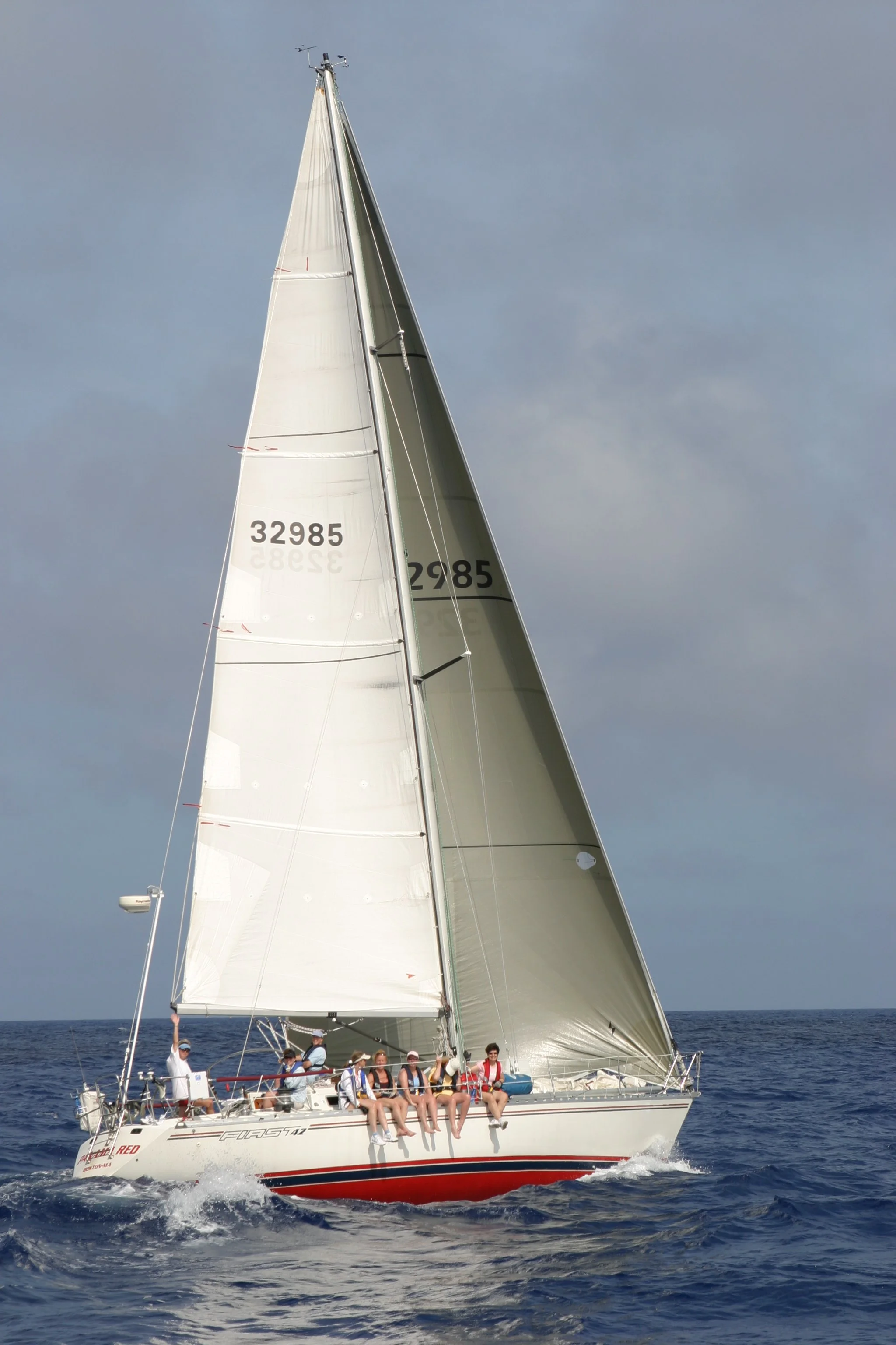 A group of people on a sailboat with white sails, sailing on the ocean under a cloudy sky.