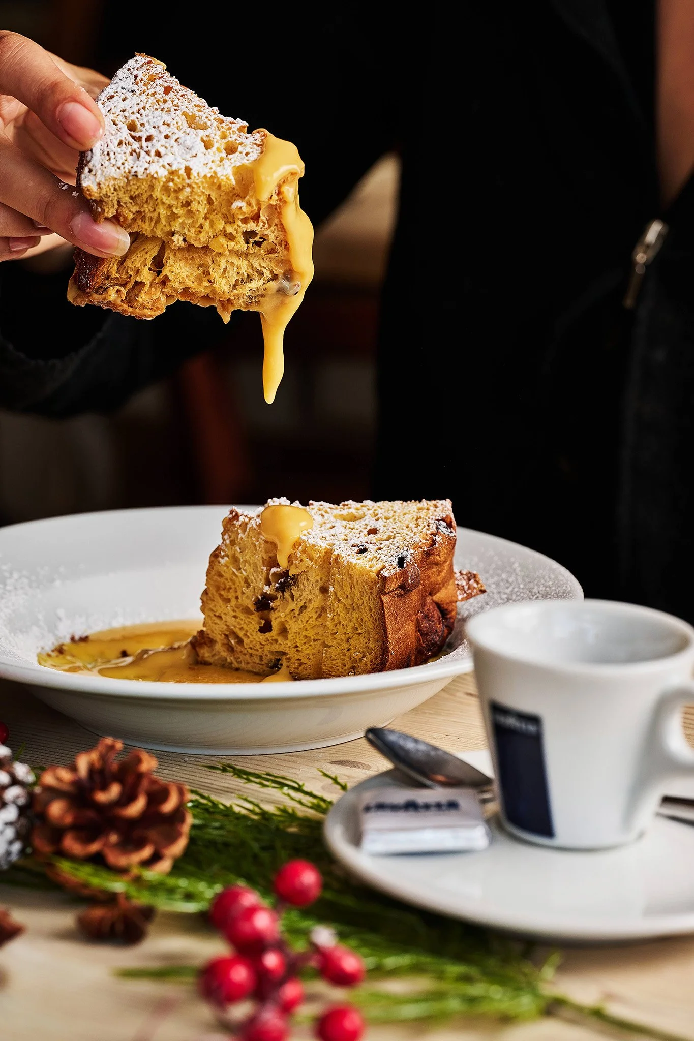 A slice of cake with powdered sugar and yellow filling being lifted from a white plate. The cake is on a wooden table, surrounded by festive decorations, including red berries and pinecones.