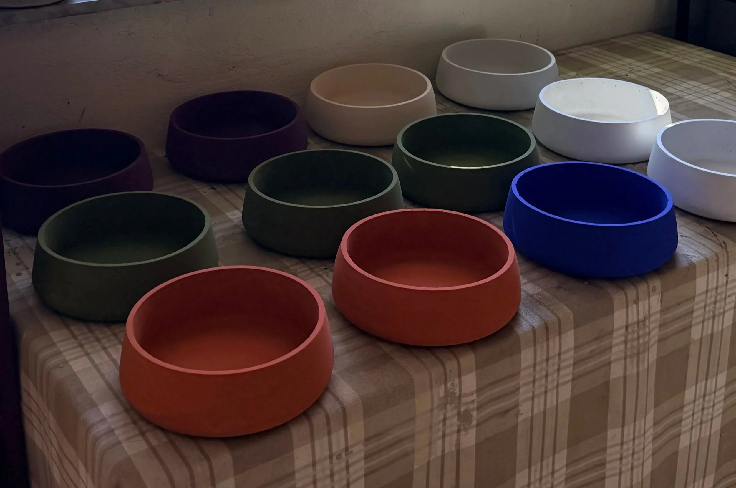 Multiple ceramic bowls in various colors including white, green, blue, purple, and red are arranged on a wooden surface with a checkered tablecloth. The background is a plain wall telling an indoor scene.