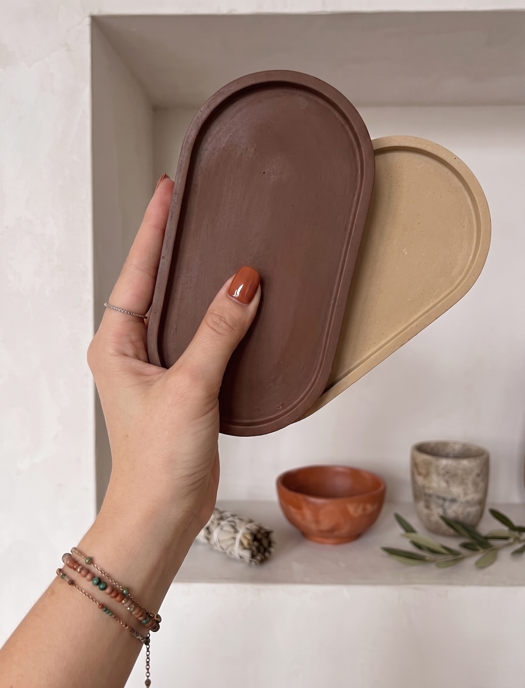 Hand holding three oval-shaped ceramic trays in earthy tones, with a shelf of various patterned and colorful bowls in the background.