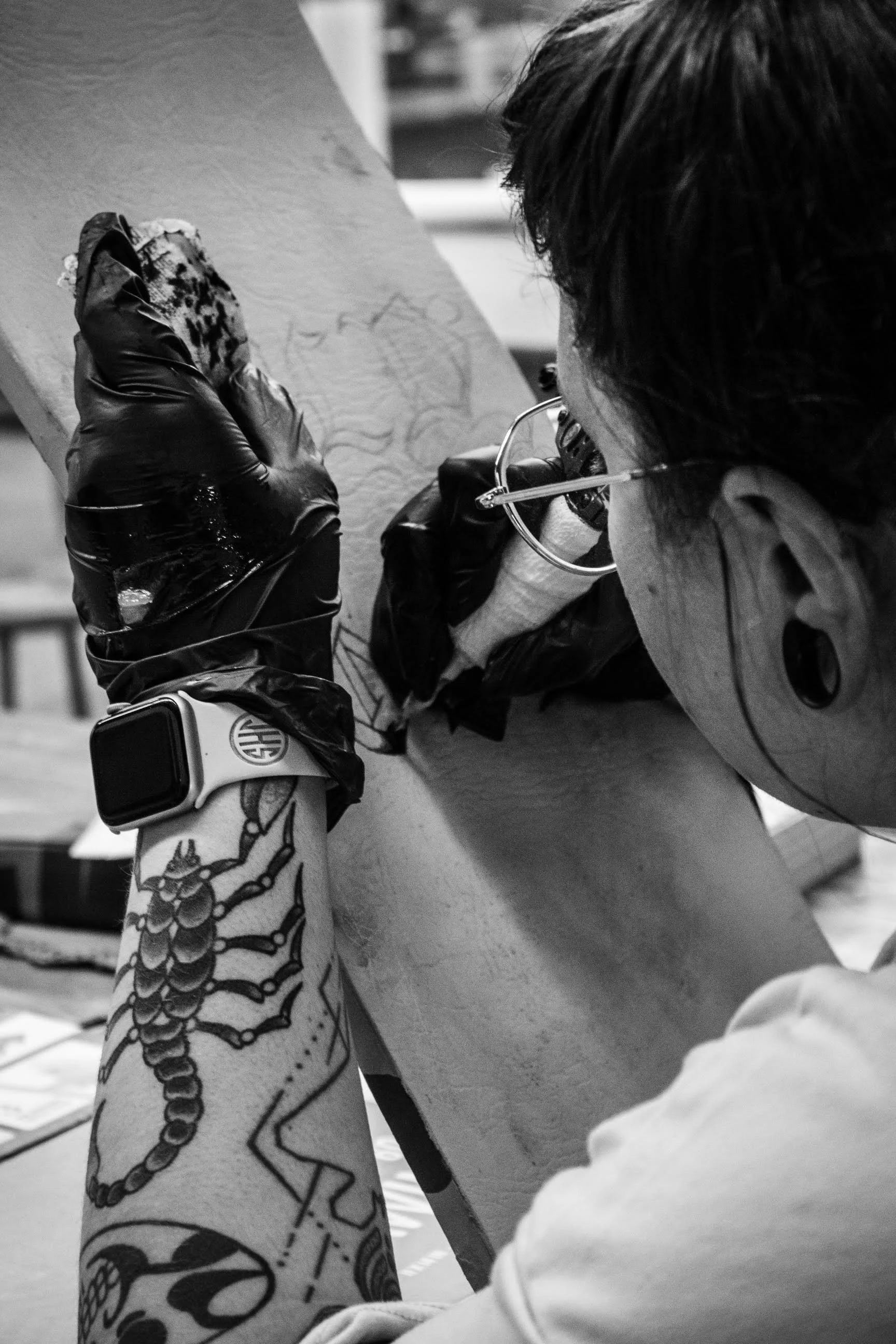 A black-and-white, close-up photograph shows an artist from a high-angle, over-the-shoulder perspective, actively tattooing a design onto a light-colored surface.
