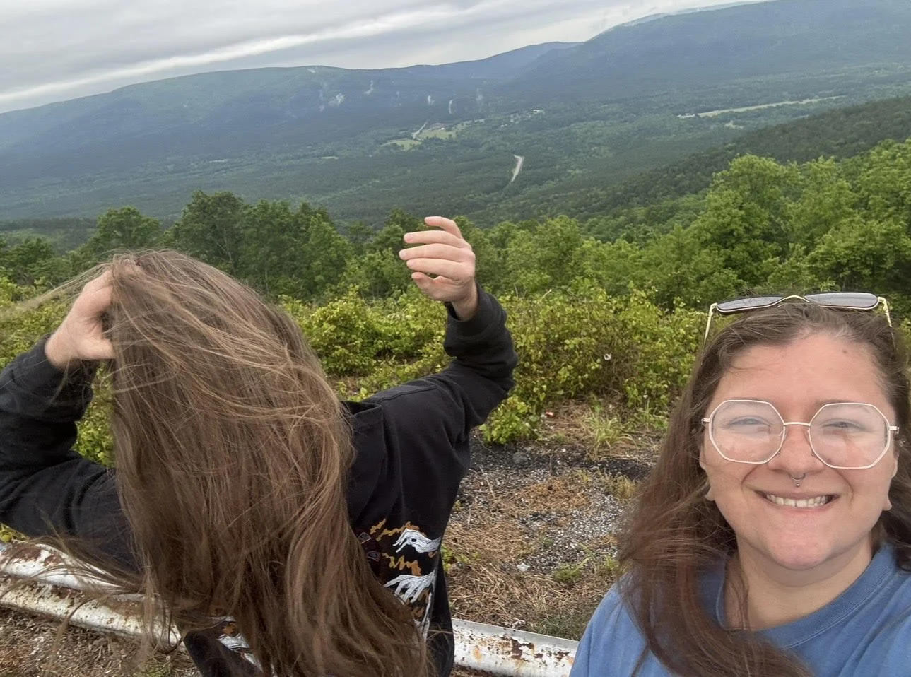 An image of Sydni, the artist, on the right side of the screen, smiling and her husband on the left side, facing away from the camera while he fixes his hair in the wind.