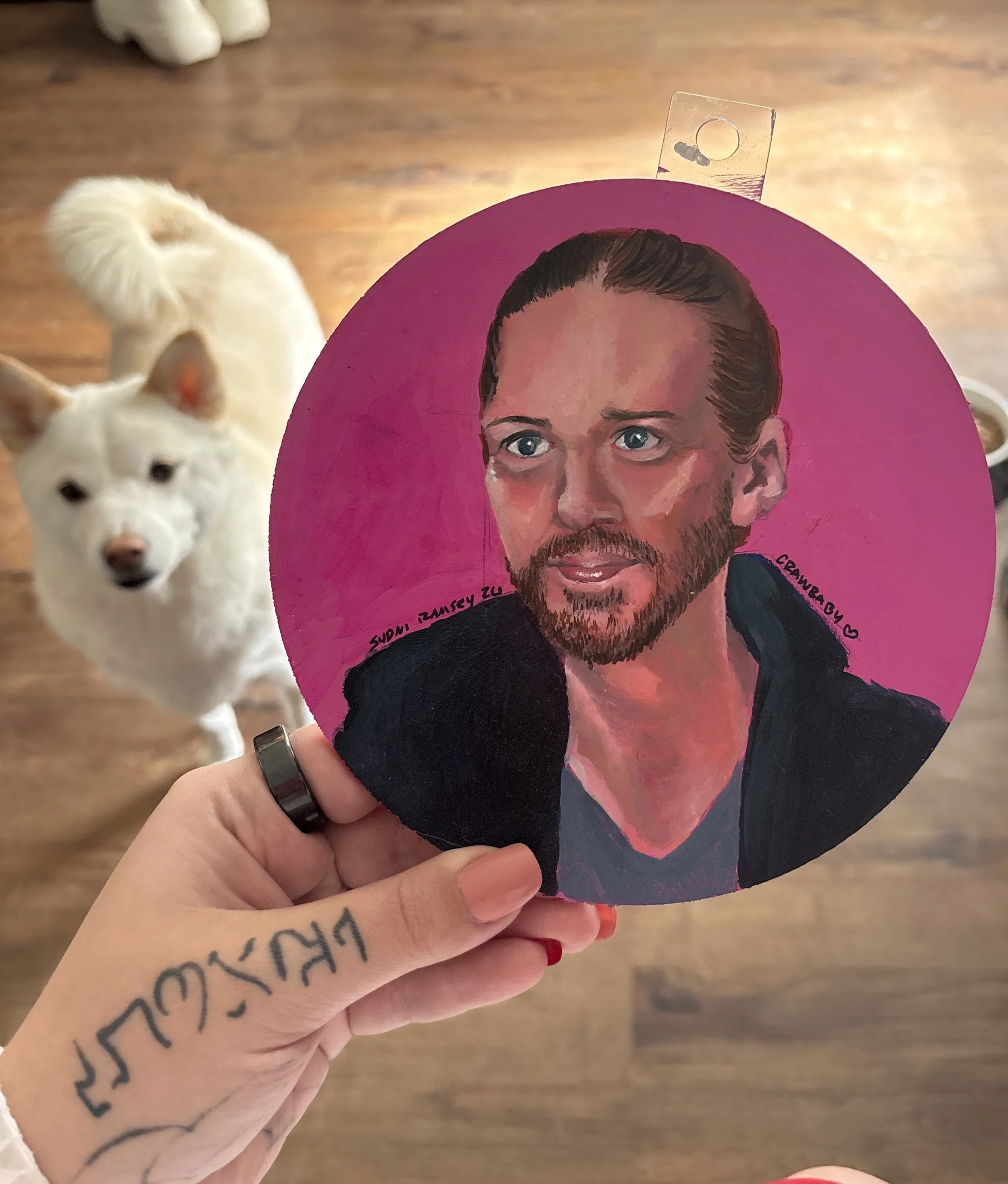 Person with a tattoo on their hand holding a pink circular card with a painted portrait of a man with dark hair, a beard, and blue eyes, against a wooden floor background. Part of a white dog with a curly tail is visible in the background.