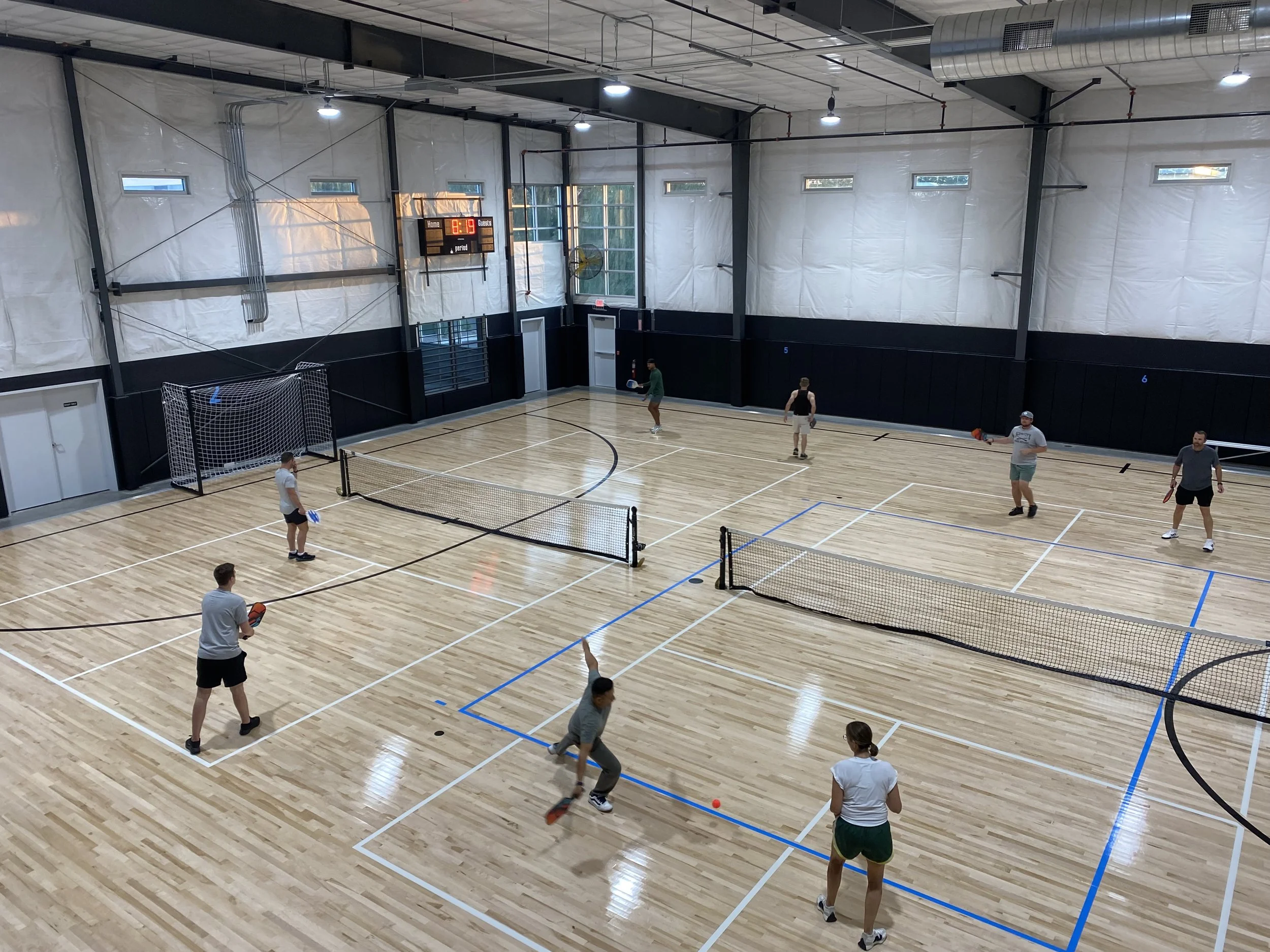 An indoor gymnasium with a wooden floor and multiple people playing pickleball. There are nets set up across the court, a scoreboard showing 1:19, and windows along the walls allowing natural light in.