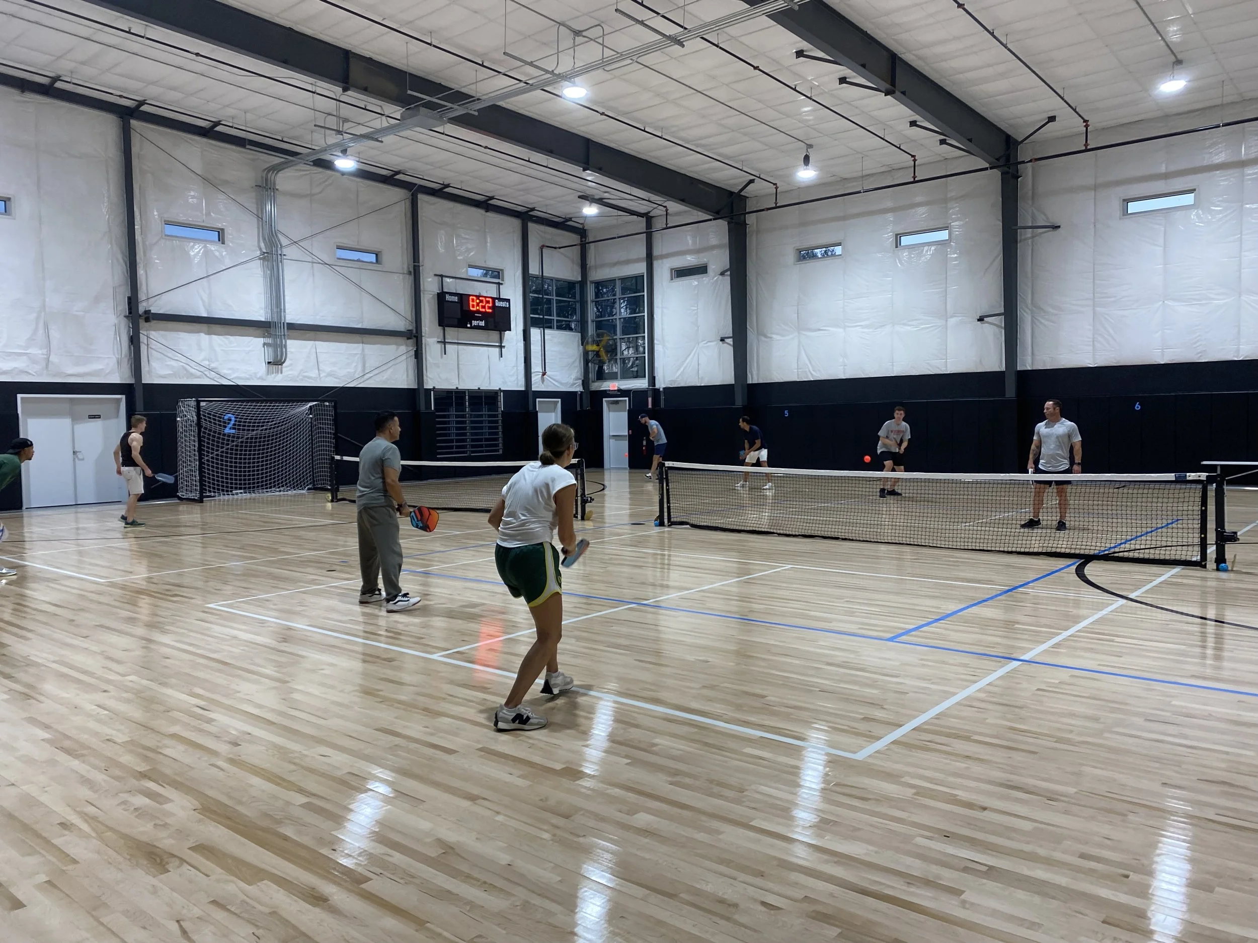 Indoor sports facility with a basketball court and a pickleball game in progress. Several players are on the court, and a digital scoreboard shows 8:22 remaining.