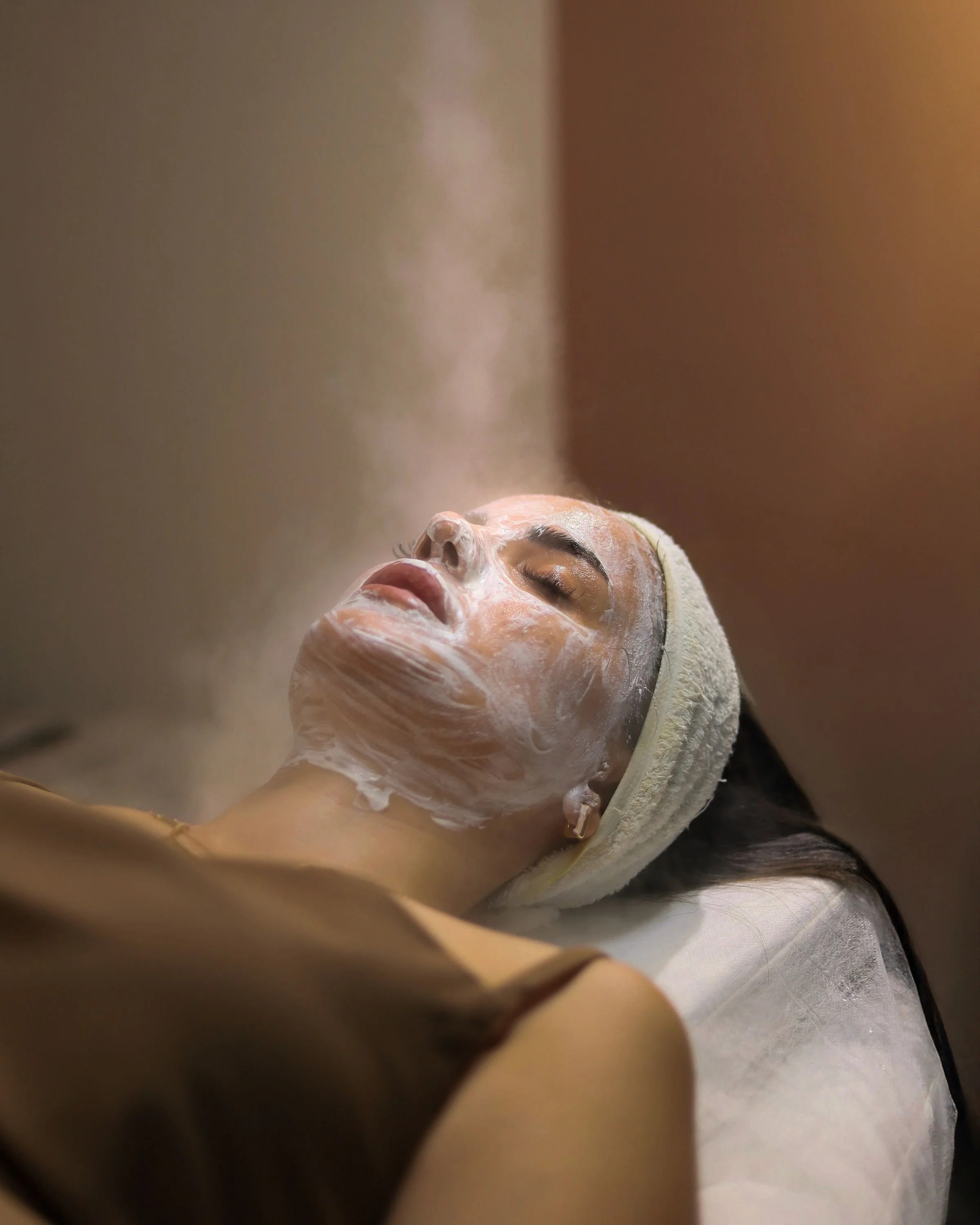 A woman lying on a treatment bed with her eyes closed, receiving a facial treatment. She has a white facial mask on her face, and a towel wrapped around her head. The image is taken indoors, with warm lighting in the background.