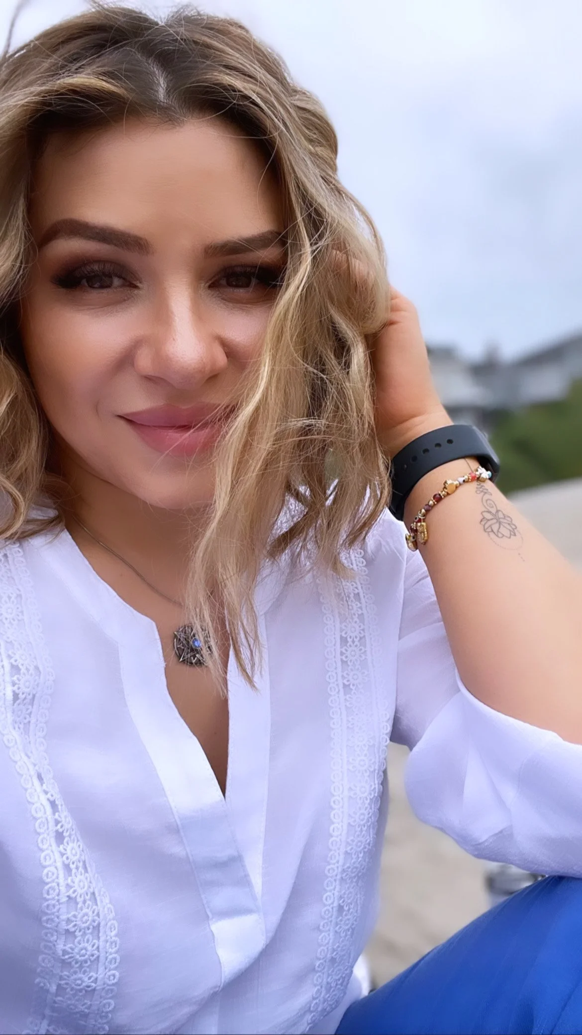 A woman with wavy blonde hair, wearing a white embroidered blouse, black wristband, and jewelry, smiling outdoors with cloudy sky and blurred background.