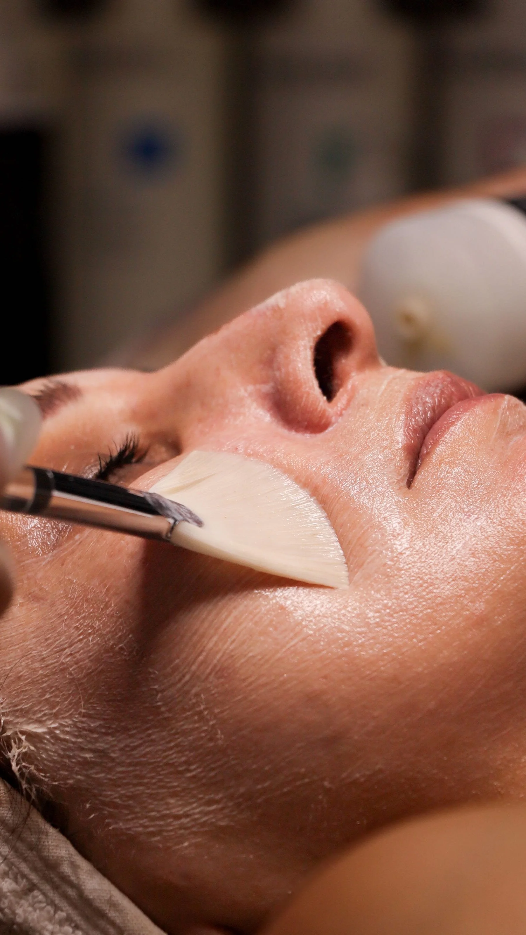 Close-up of a person receiving a facial treatment, with a brush applying a skincare product to their face.