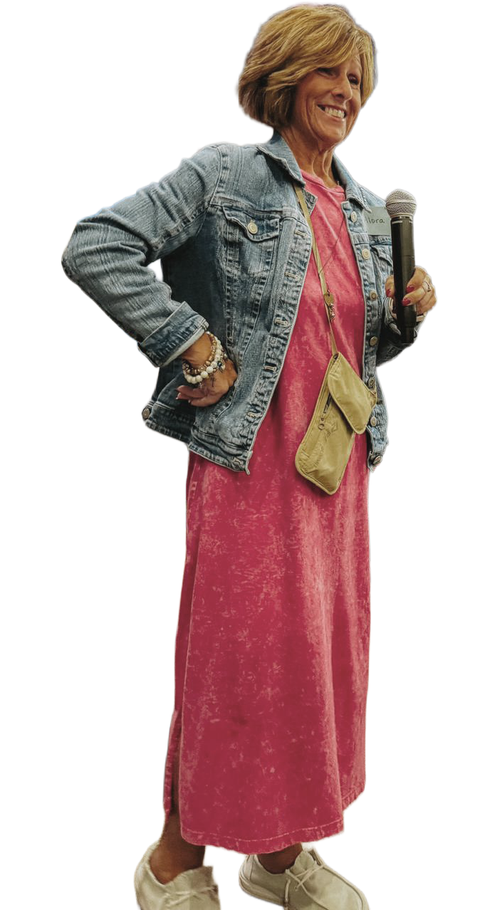 Alora Hosfield dressed in a pink maxi dress and denim jacket, holding a microphone, standing with one hand on her hip and smiling.