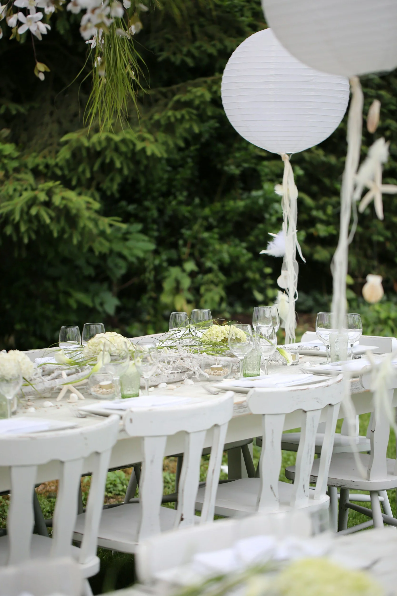 An outdoor white dining table decorated with white flowers, glassware, and lanterns, set for a gathering, surrounded by green foliage and trees, with white paper lantern lights hanging above.