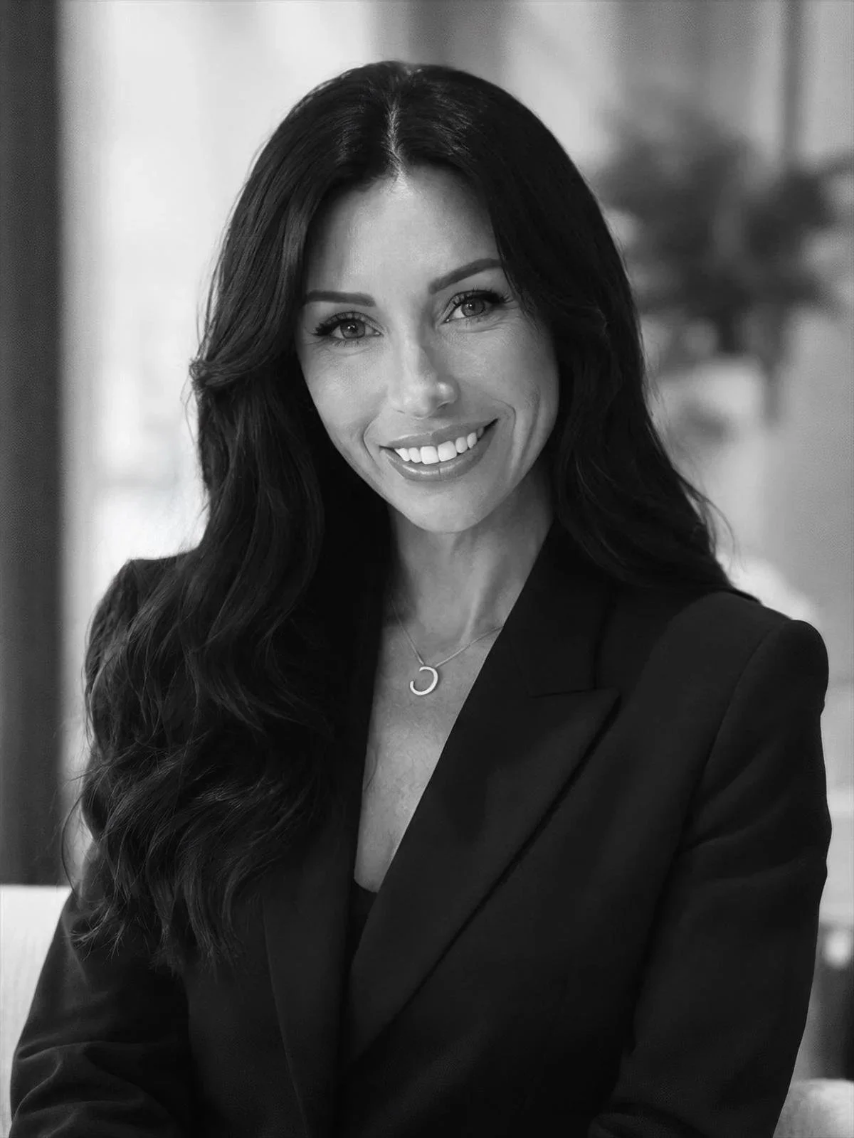 A smiling woman with long dark wavy hair, wearing a black blazer and a necklace with a crescent moon pendant, in an indoor setting.