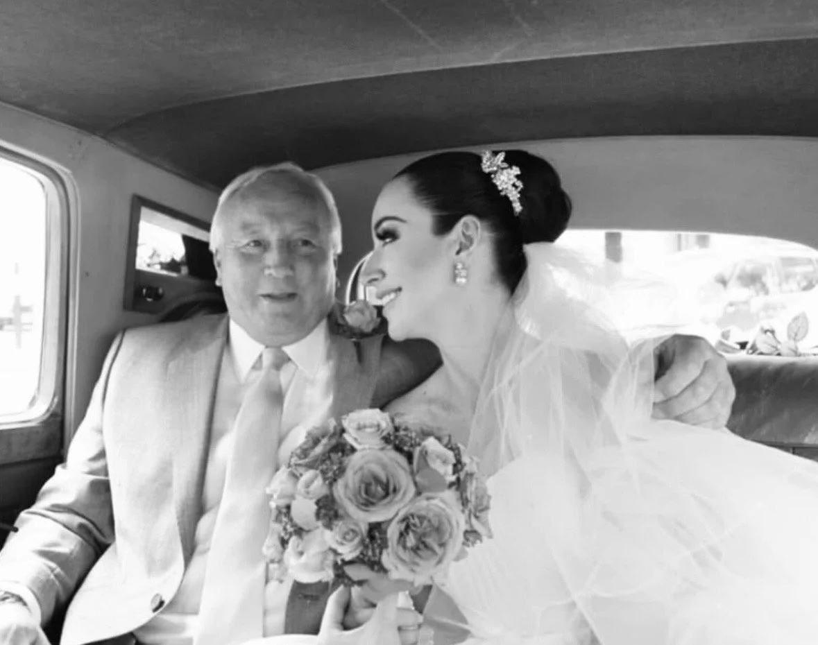 Black and white photo of a bride and an older man in a vehicle with the bride holding a bouquet of roses, sharing a joyful moment.