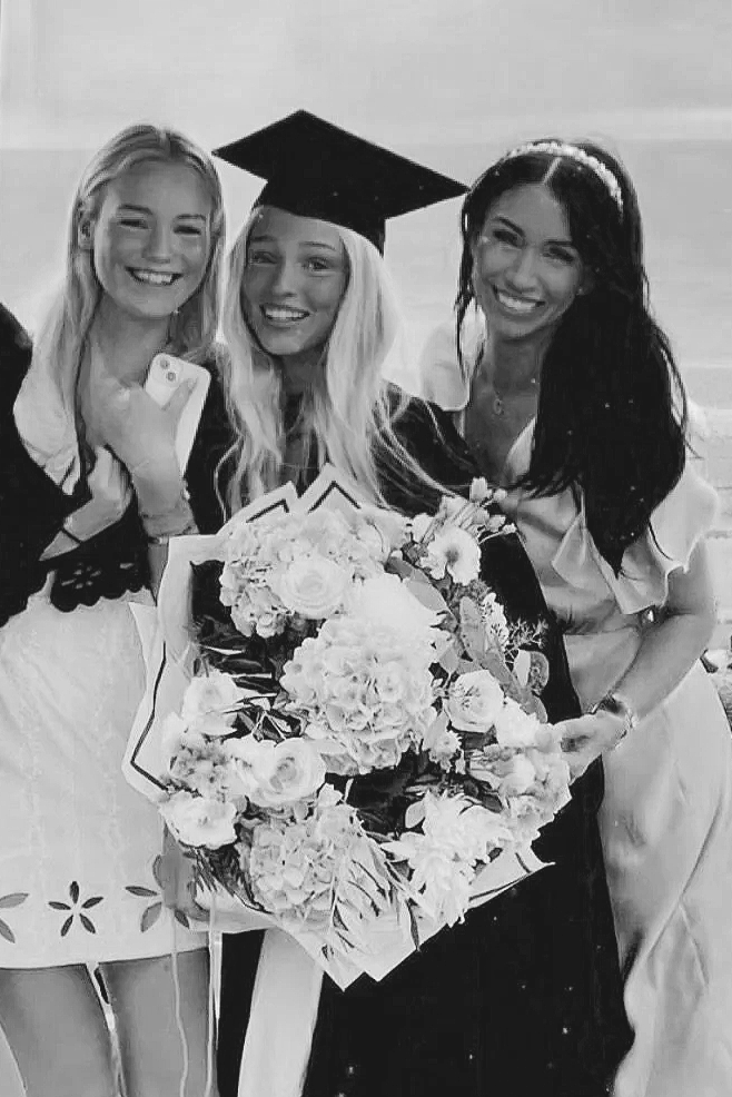 Three women celebrating graduation, with one woman in a graduation cap and gown holding a large bouquet of flowers, smiling