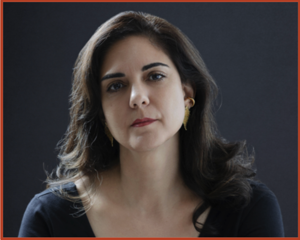 Portrait of a woman with long dark hair, wearing gold earrings and a black top, looking serious against a dark background.