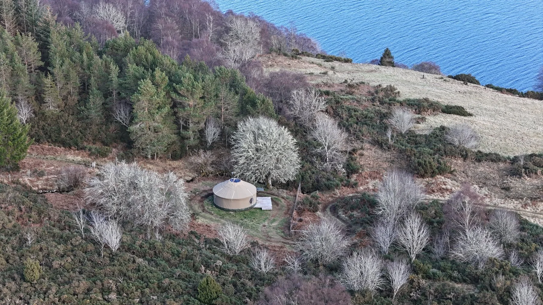 A yurt situated in a rural retreat area, in a hilly autumn landscape with trees and shrubs, overlooking Loch Ness in the background.