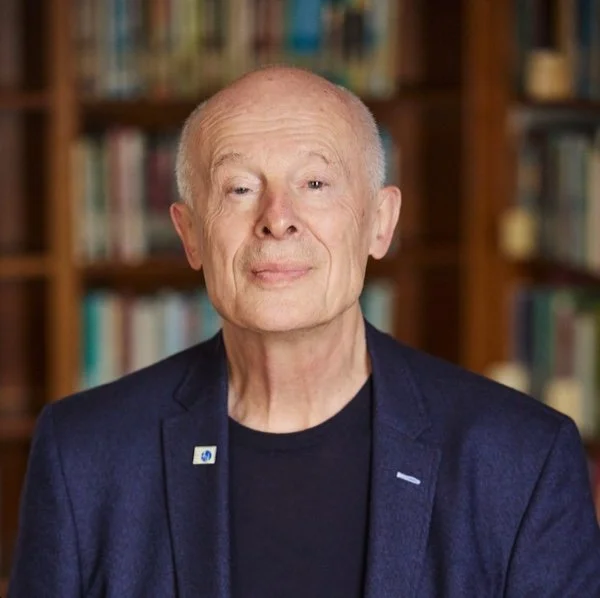 John Schellnhuber, Director General of IIASA. A senior man with a slight smile, wearing a dark blazer and black shirt, sitting in front of a bookshelf filled with books.