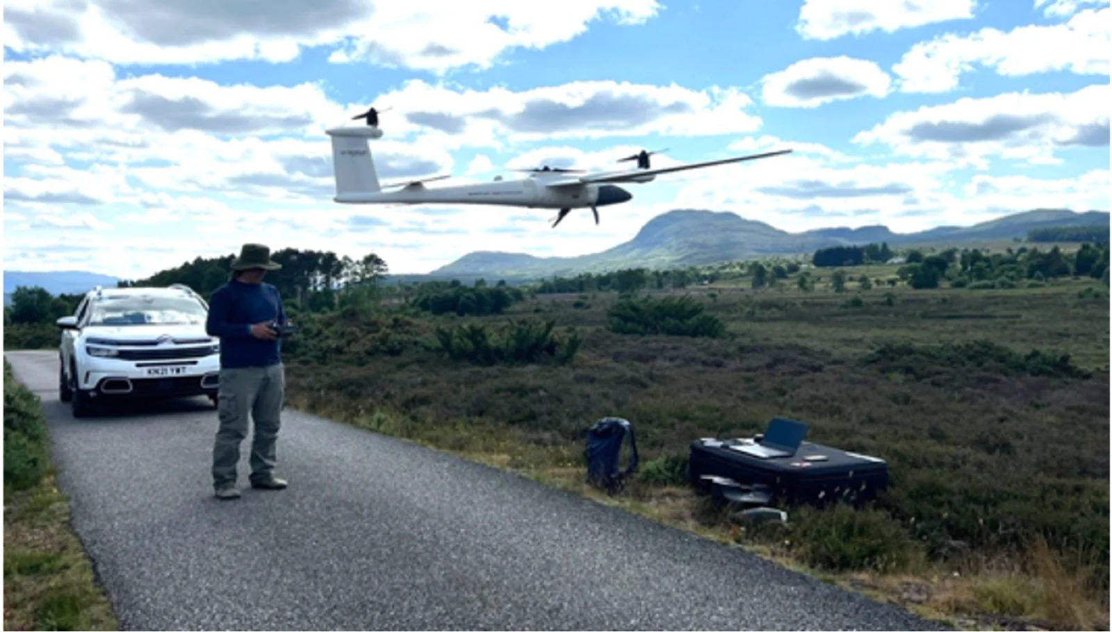 A man in a hat operating a drone with a surveying drone flying overhead, conducting biodiveristy surveys. A white car parked on a rural road, and a laptop set on a backpack in a scenic countryside with mountains and a partly cloudy sky.