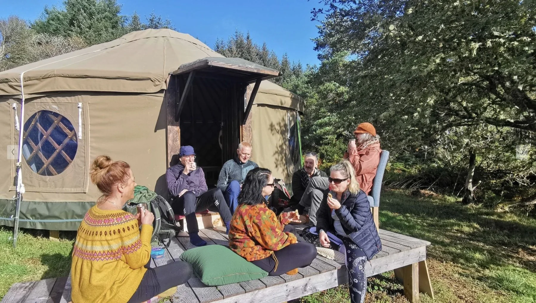 A group of eight people gathered outside a large yurt, sitting on a wooden platform with some cushions. They are enjoying food and drinks on a sunny day, surrounded by trees and greenery.