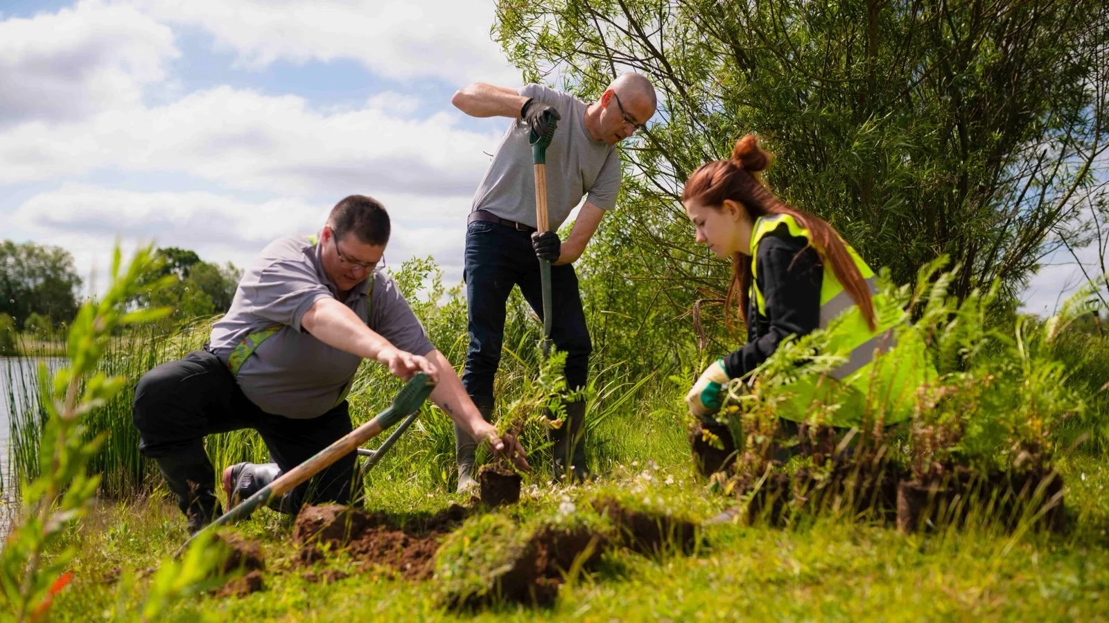 Three people volunteering in nature, planting a tree by a lochan under a cloudy sky, two men and one woman, with the woman wearing a high-visibility vest.