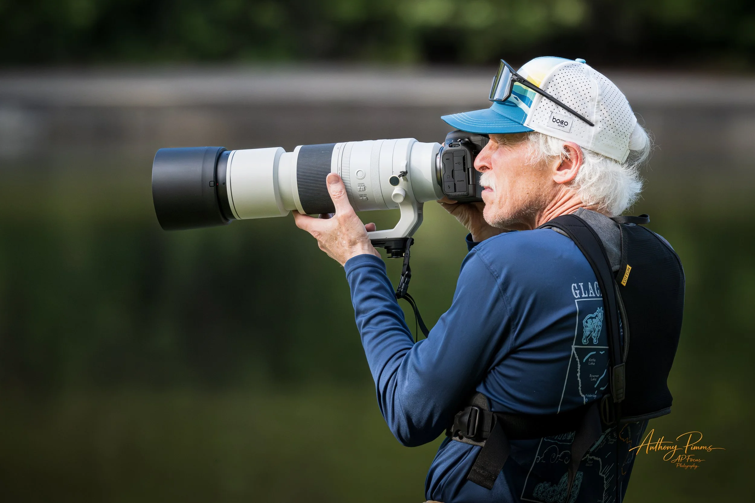 An older man with white hair wearing a blue jacket and a white and blue cap, using a large camera with a telephoto lens outdoors.