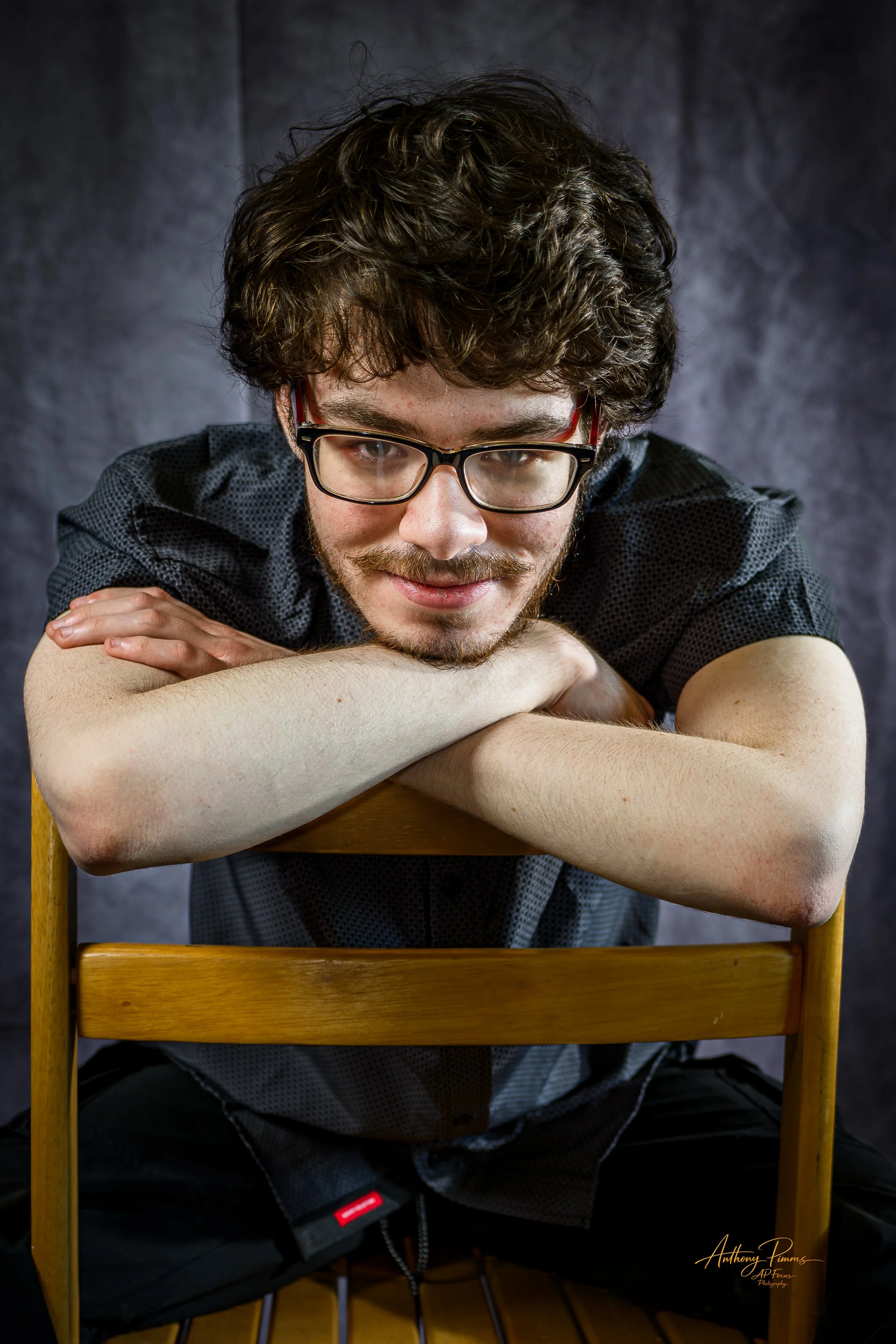 A man with glasses and curly hair leaning on a wooden chair, smiling, against a dark background.