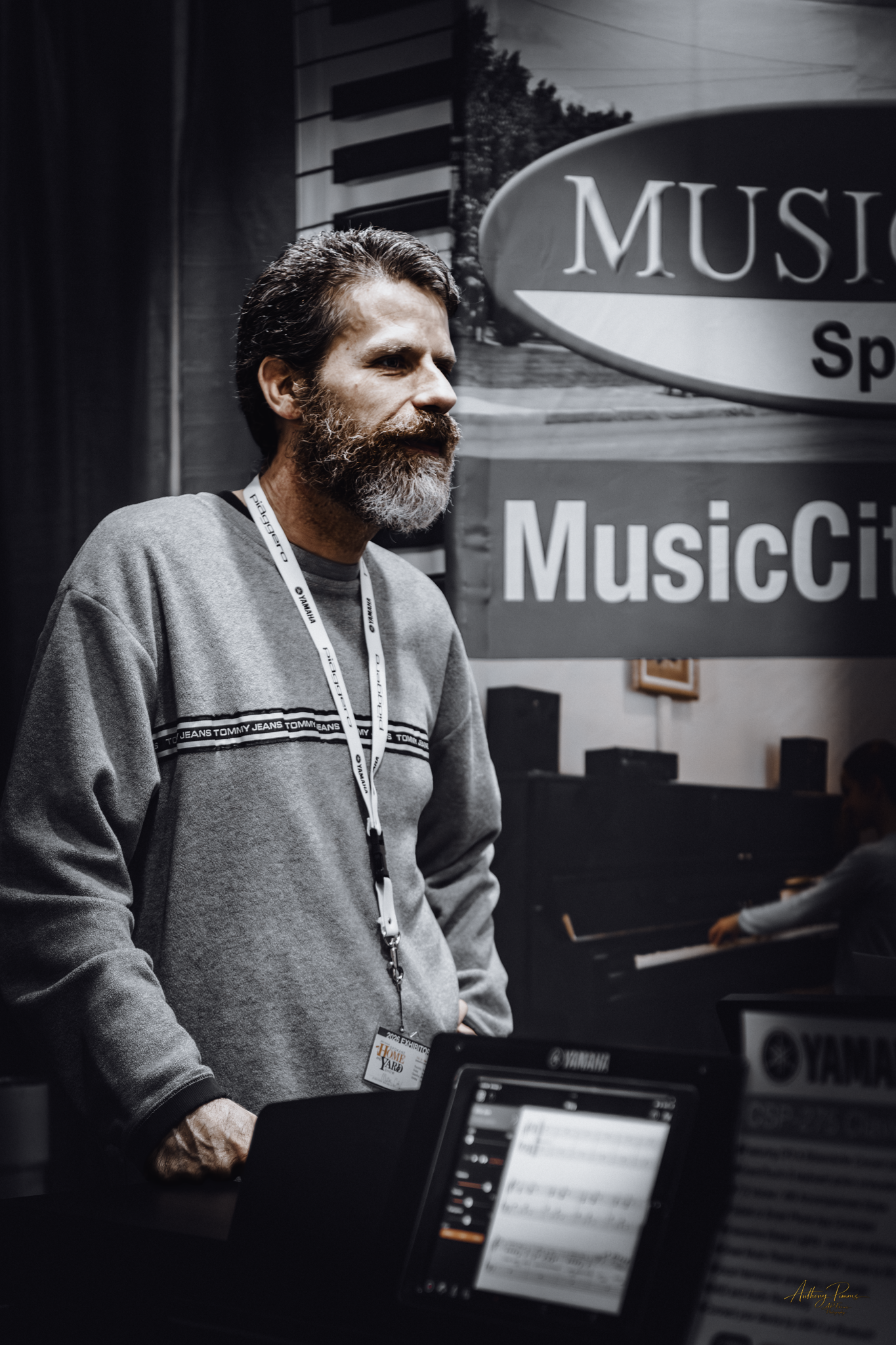 A man with a beard and dark hair standing at a music booth, with large signs reading 'Music' and 'Music City' behind him. There are electronic music equipment and laptops in front of him.