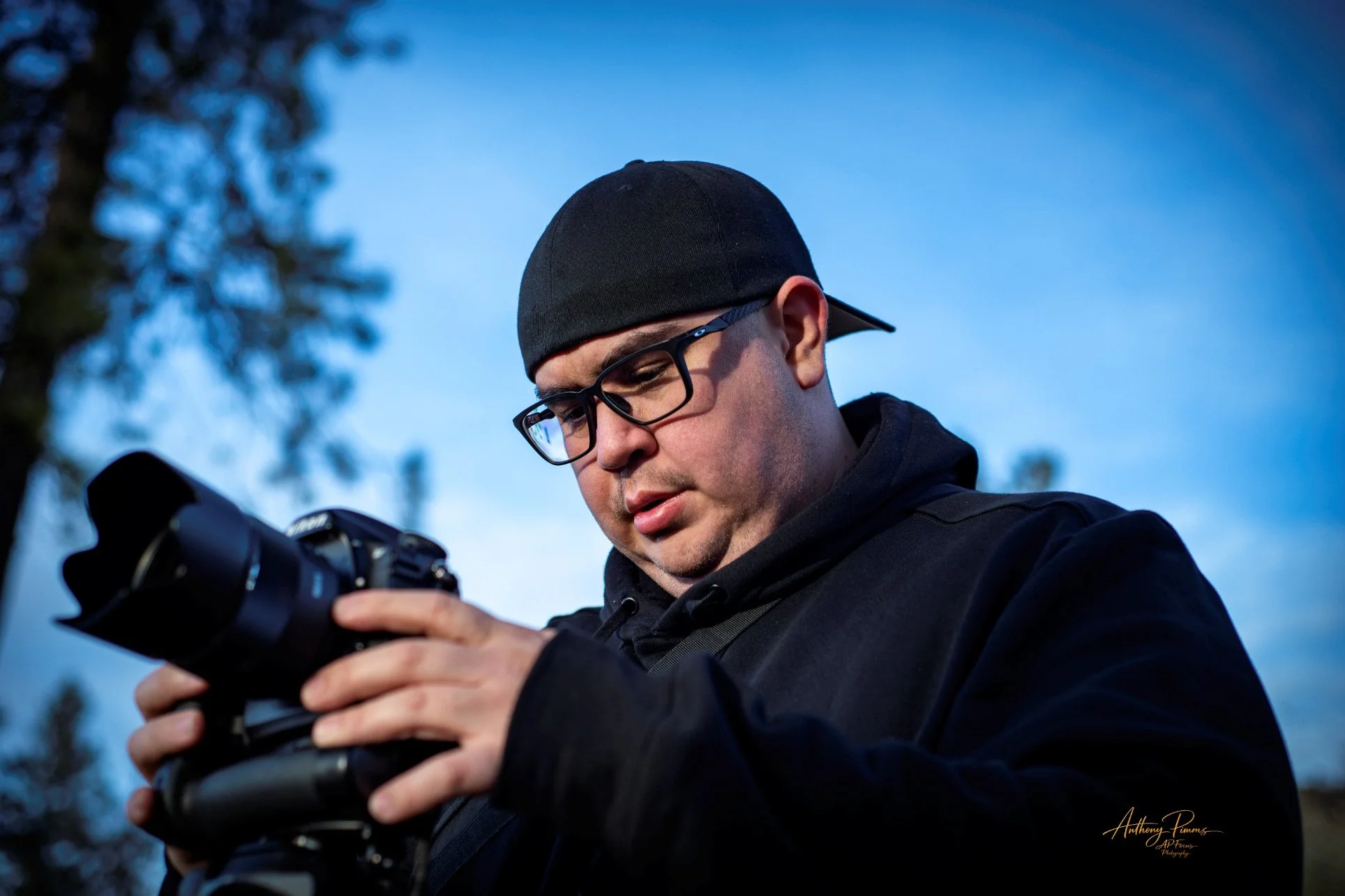 A man wearing a black cap, black glasses, and a black hoodie is looking at a camera he is holding in his hands outdoors during the daytime. There are blurred trees and a blue sky in the background.
