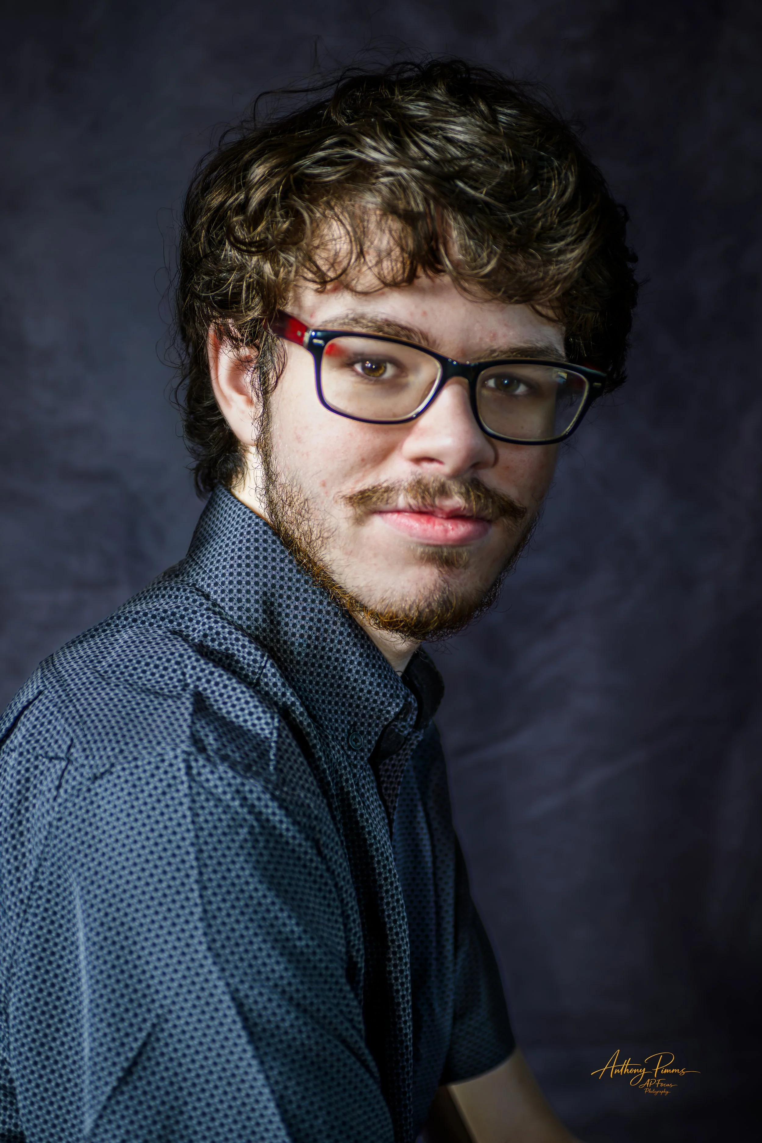 Young man with curly brown hair and a beard, wearing black-rimmed glasses and a patterned button-up shirt, posing against a dark textured background.
