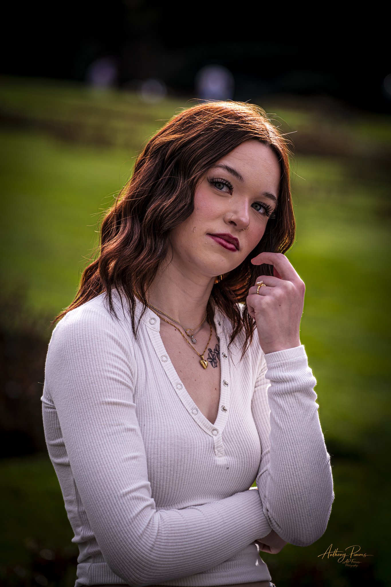 A young woman with wavy reddish-brown hair and blue eyes posing outdoors against a blurred green background, wearing a white long-sleeve shirt, necklace, and ring, with sunlight highlighting her hair.