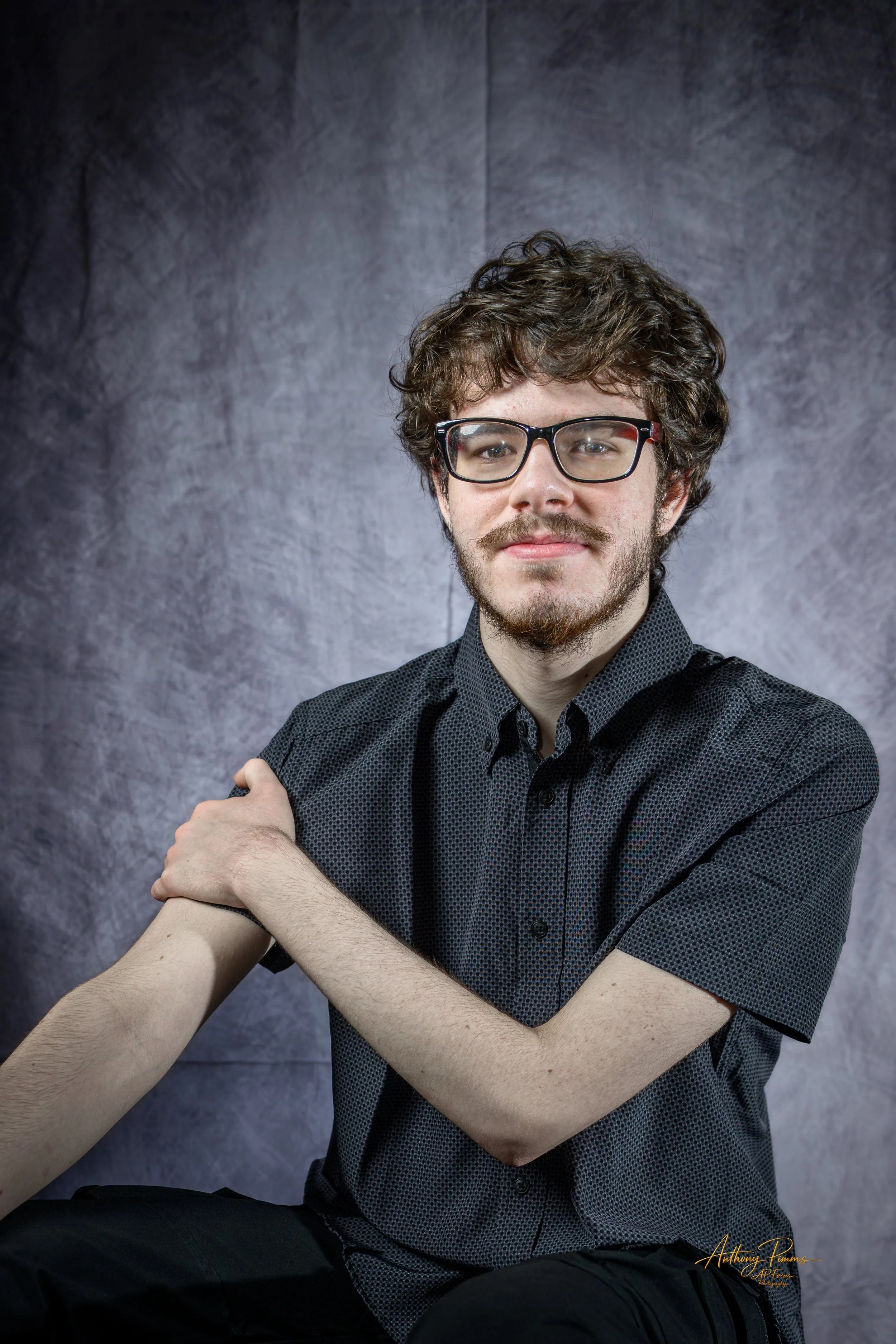 A young man with curly hair, glasses, a beard, and mustache in a black dotted shirt, sitting and flexing his right arm, with a gray textured background.
