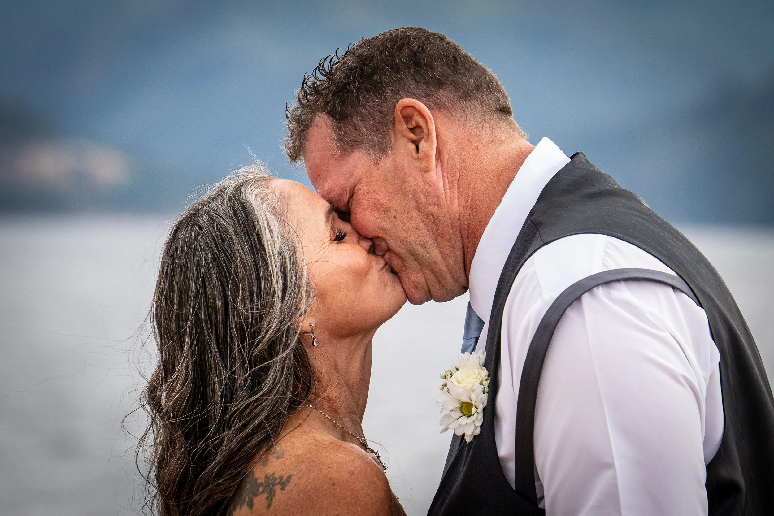 A man and woman are sharing a kiss outdoors during a wedding, with the man dressed in formal attire and the woman showing her tattooed shoulder.
