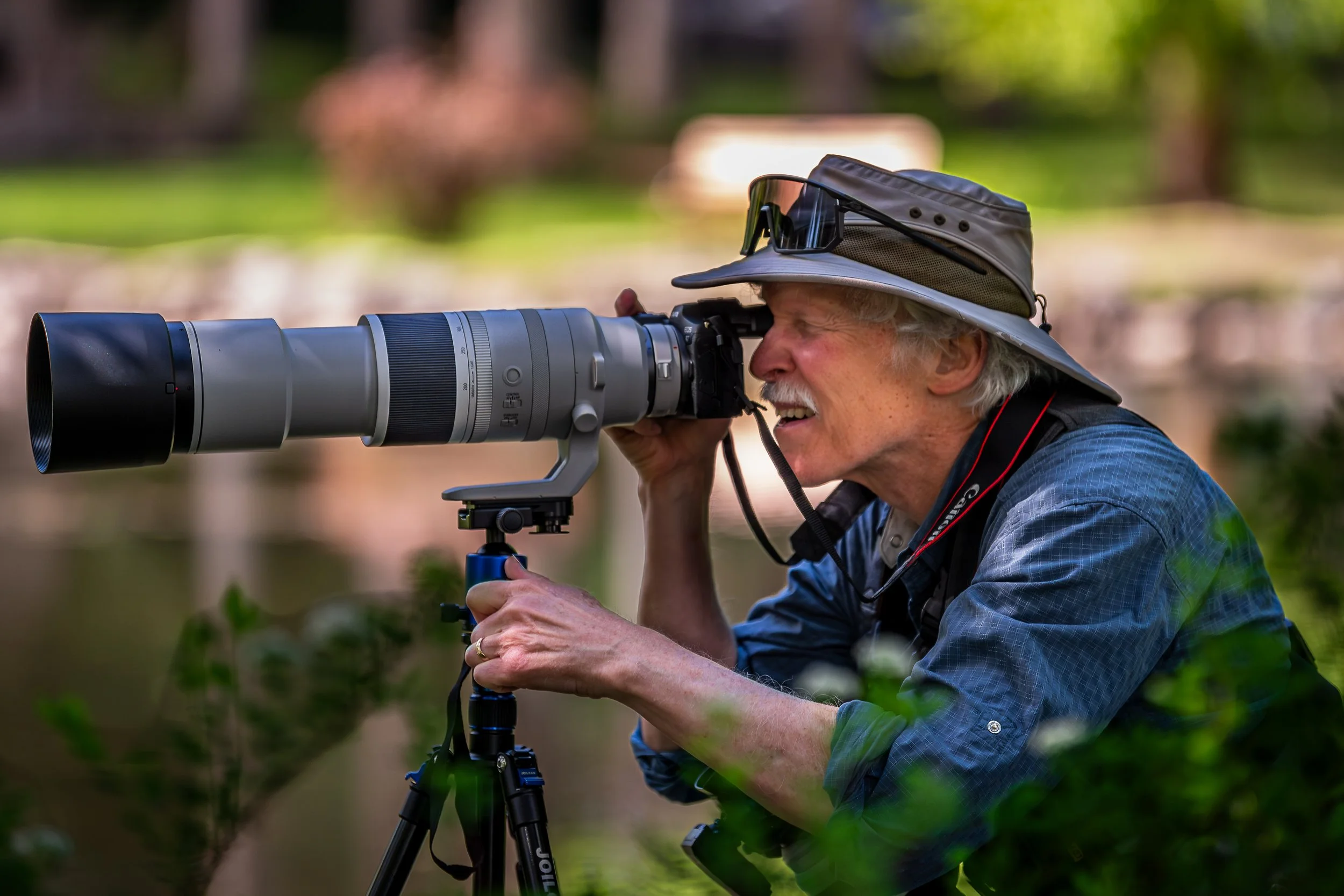 An elderly man wearing a wide-brimmed hat and sunglasses, using a large camera with a telephoto lens mounted on a tripod, outdoors in a natural setting with trees and greenery.