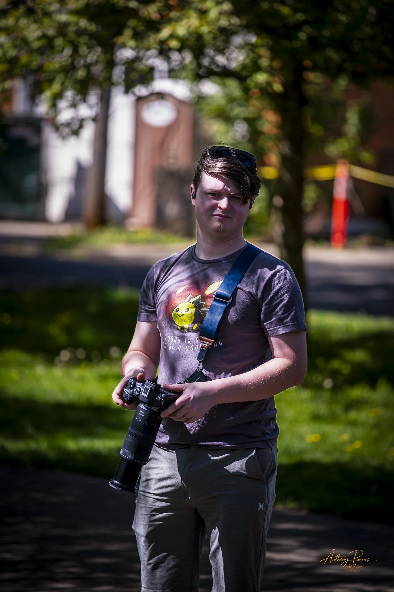 A young man with sunglasses on his head holding a camera outdoors on a sunny day, with a blurred background of trees and buildings.