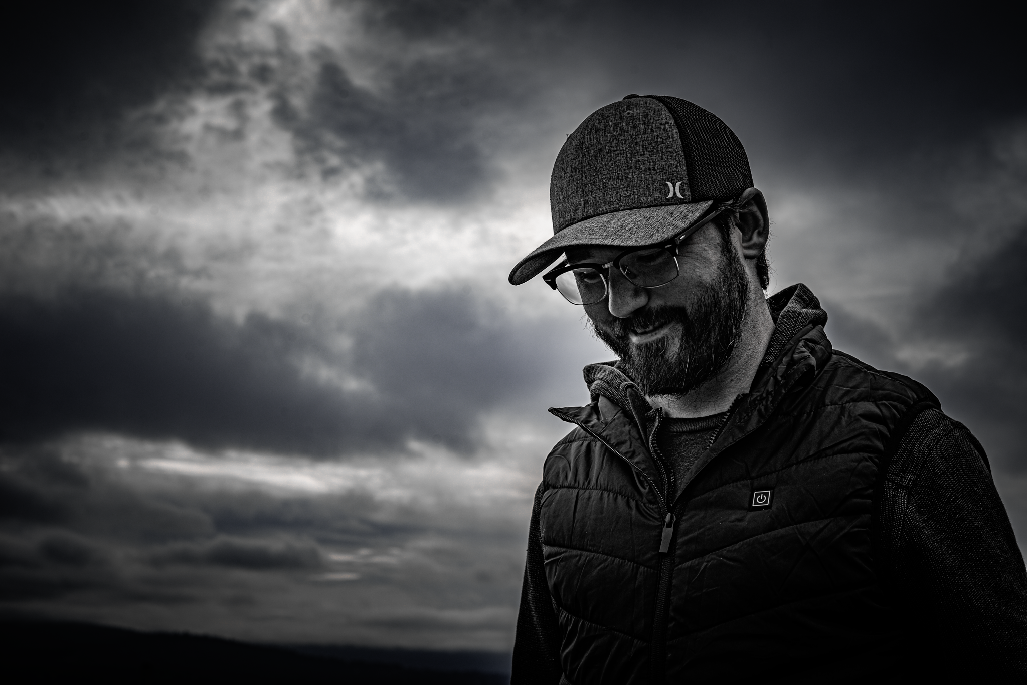 A man with glasses, a beard, and a cap looking down, wearing a winter jacket against a cloudy sky in black and white.