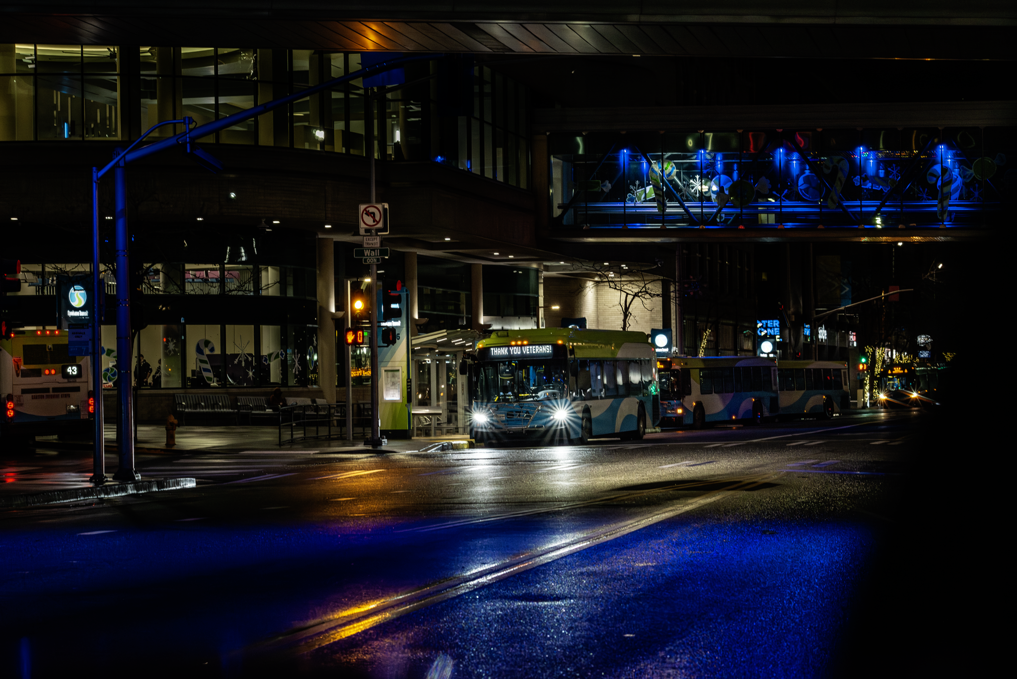 Nighttime scene showing buses at a bus stop in front of a modern building with glass windows illuminated from inside. Blue neon lights decorate the building, and there are street signs, traffic signals, and reflections on the wet pavement.