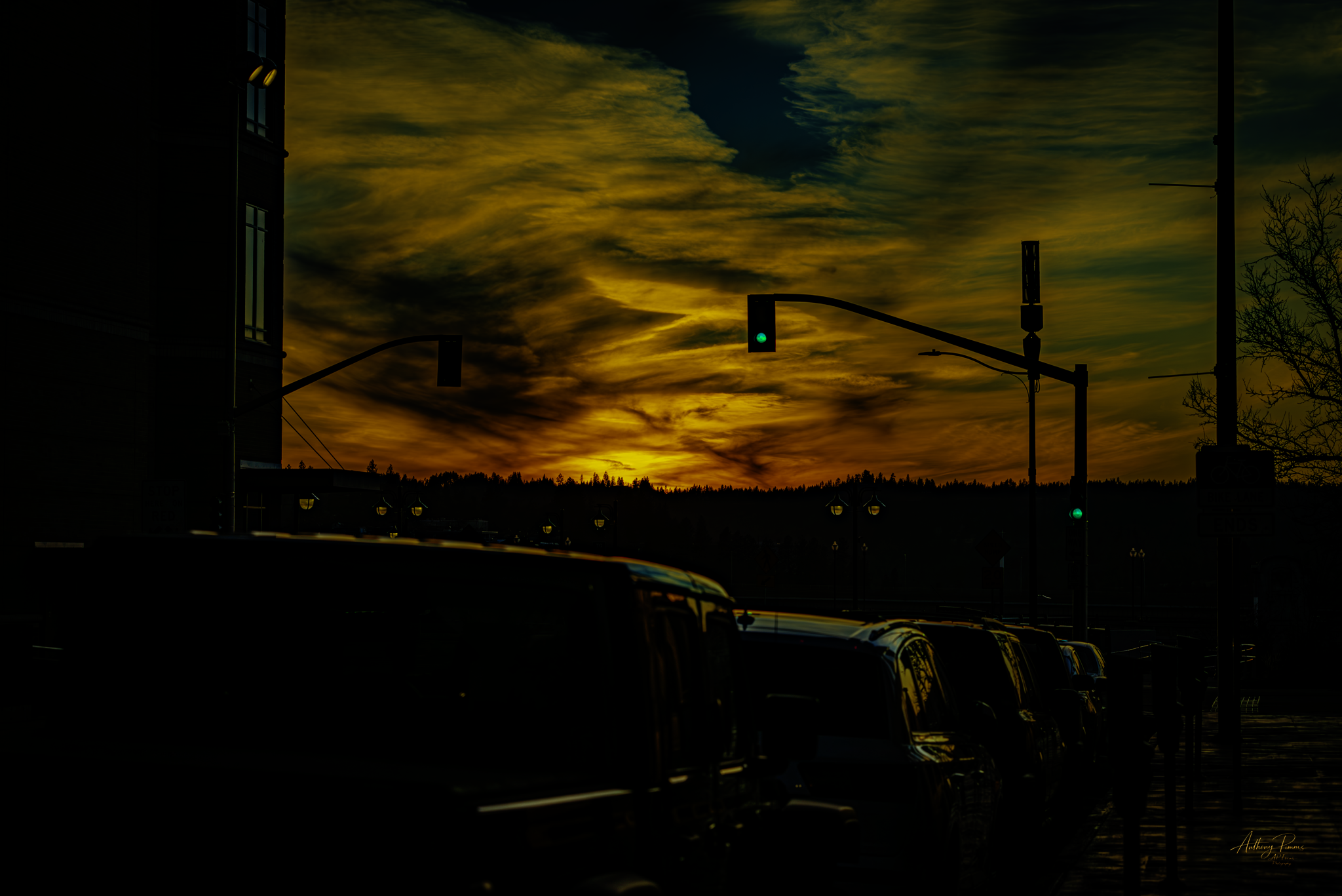 A sunset or dusk scene with a cloudy sky and the sun setting on the horizon. Silhouettes of vehicles parked along the street and traffic lights are visible, with a building on the left and some trees on the right.