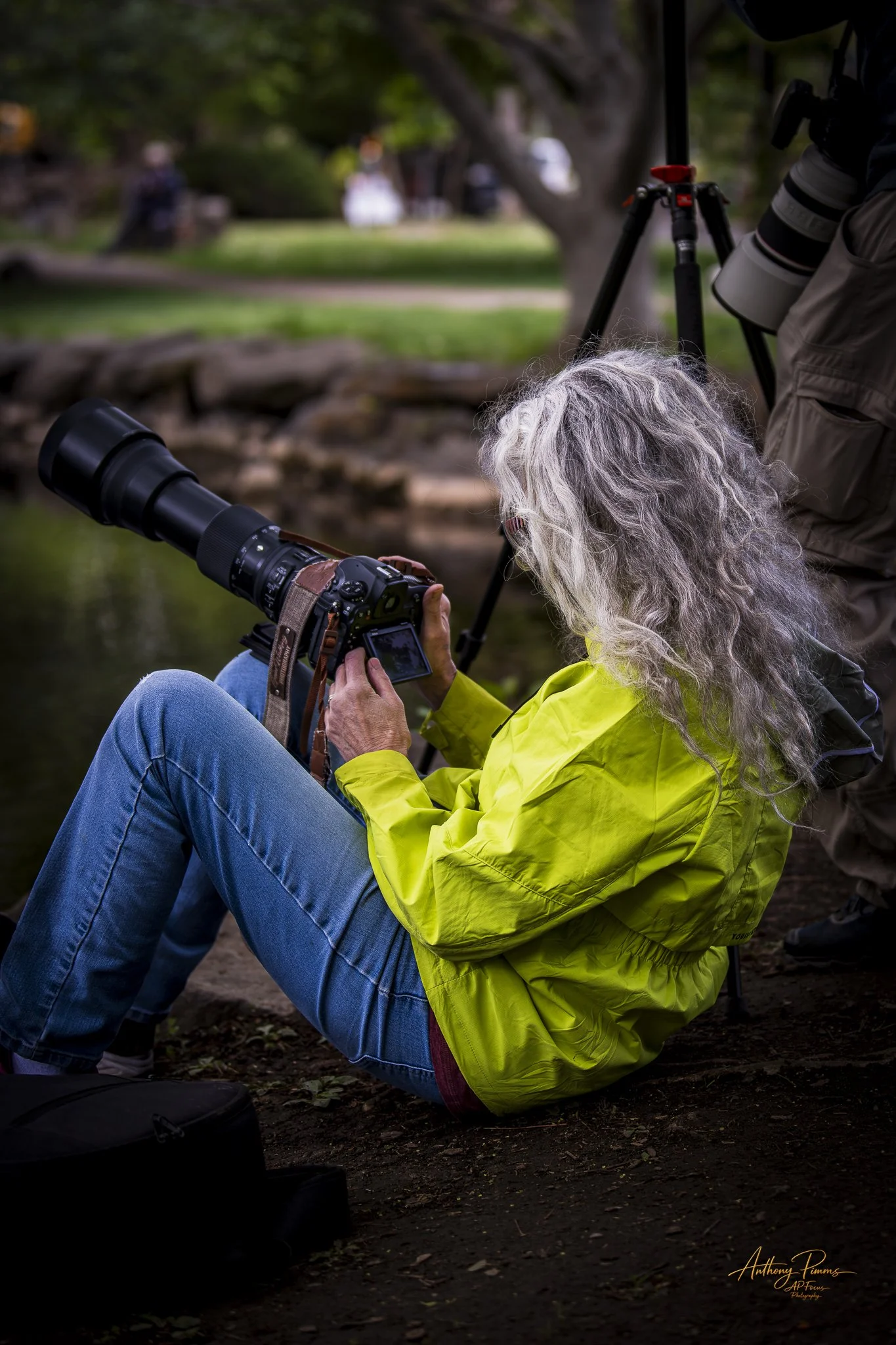 A woman with long gray hair sitting on the ground outdoors, looking at a camera with a large telephoto lens, and a person standing nearby with a camera on a tripod, in a park or garden setting.