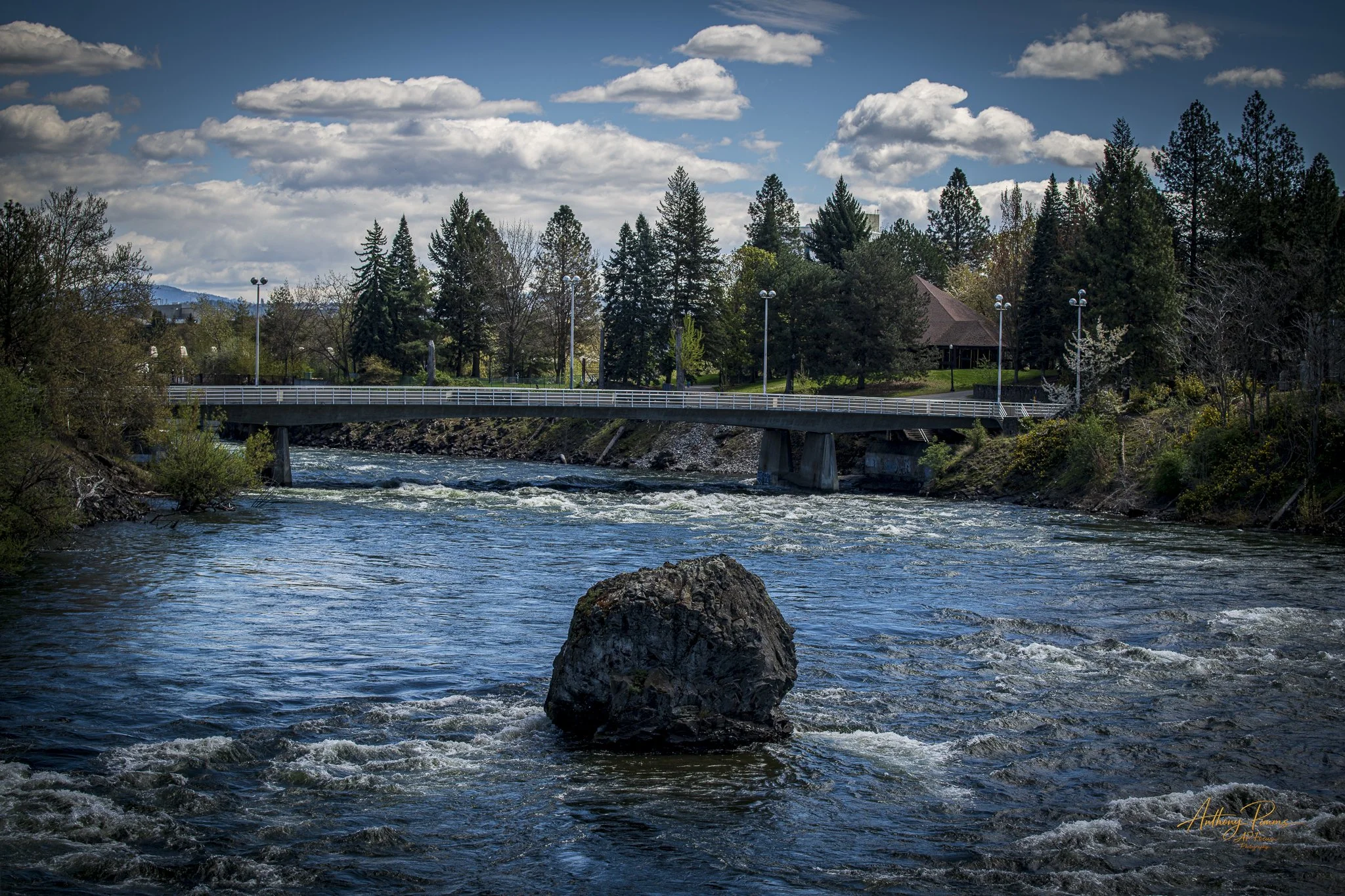 Rushing river with a large rock in the foreground and a bridge with tall trees and a building in the background under a blue sky with scattered clouds.
