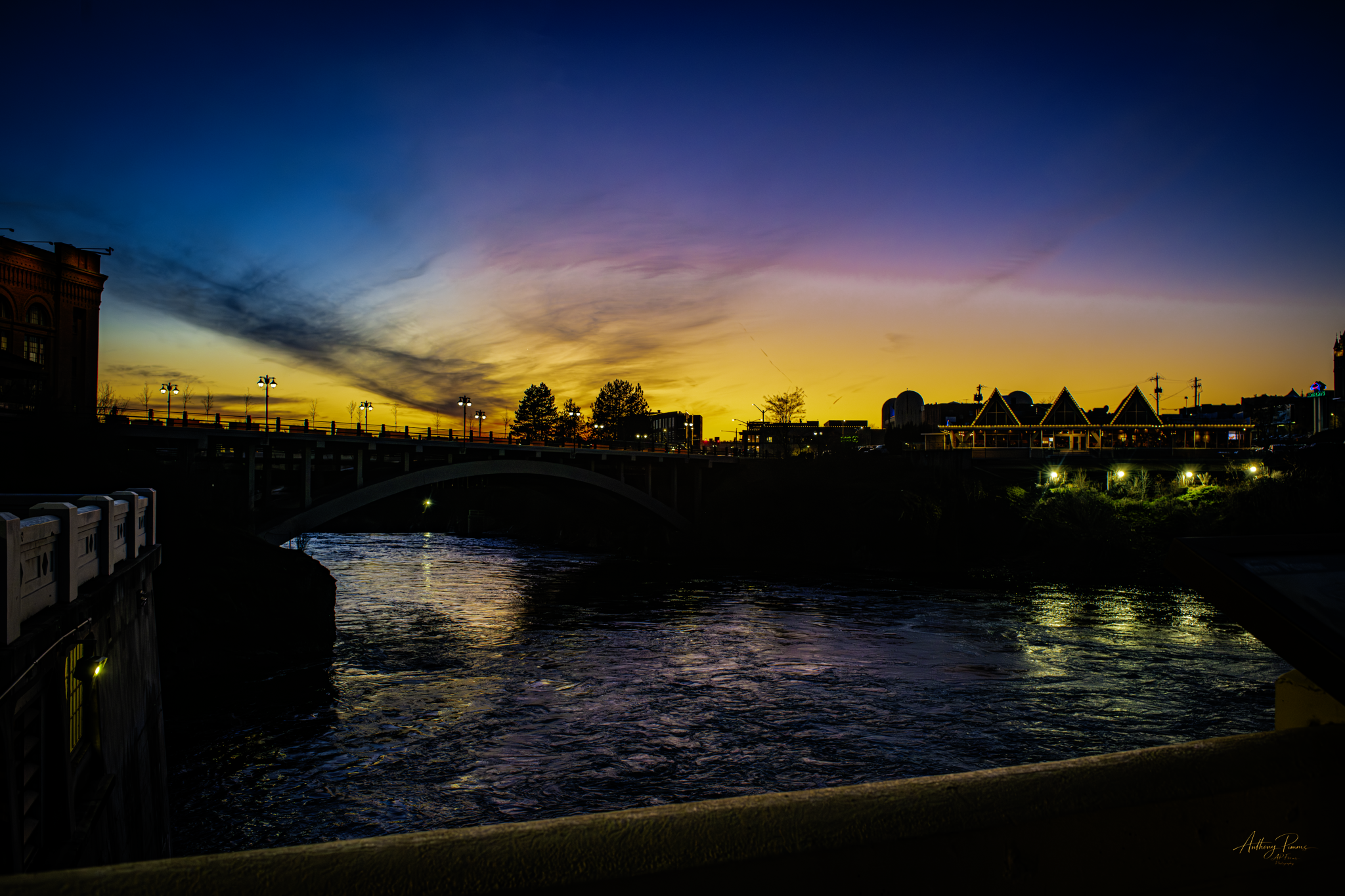Sunset scenery over a river with silhouettes of trees, buildings, and a bridge, illuminated by streetlights and fading sunlight in the sky.