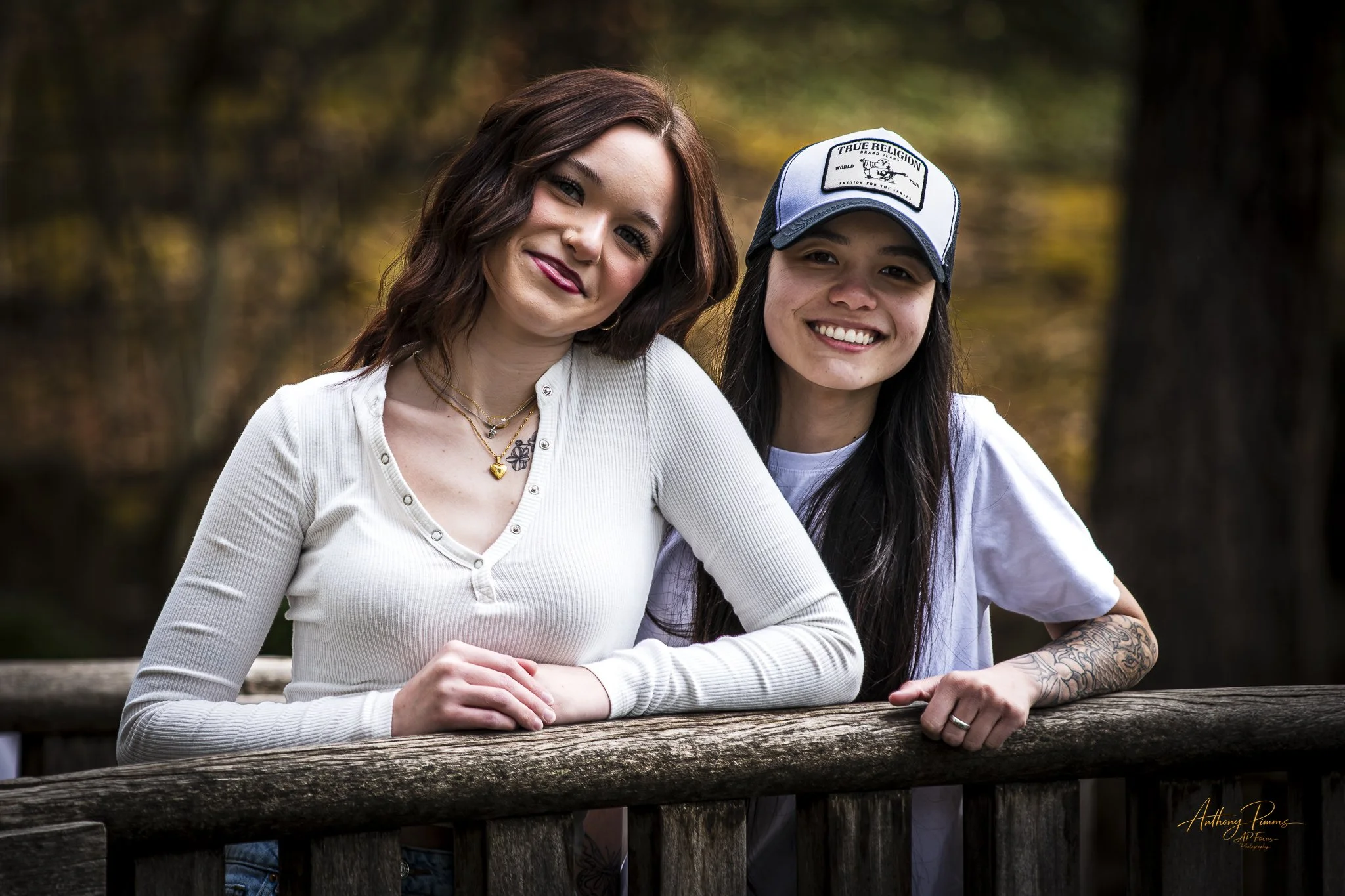 Two women posing outdoors, leaning on a wooden railing, smiling at the camera, with a blurred natural background of trees in autumn colors.