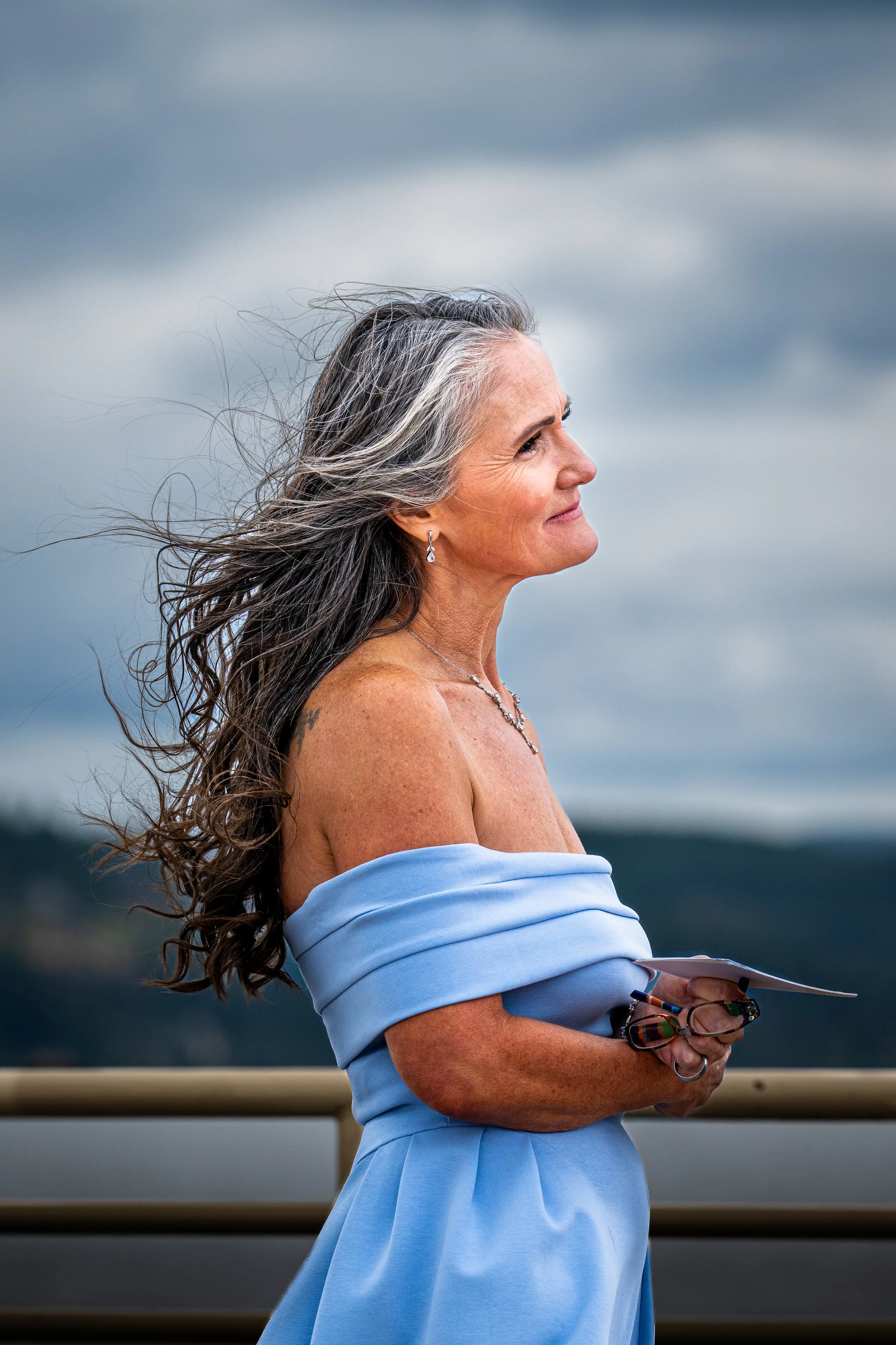 A mature woman with long, gray hair standing outdoors against a cloudy sky, wearing an off-shoulder light blue dress, holding a pair of eyeglasses and a notebook.