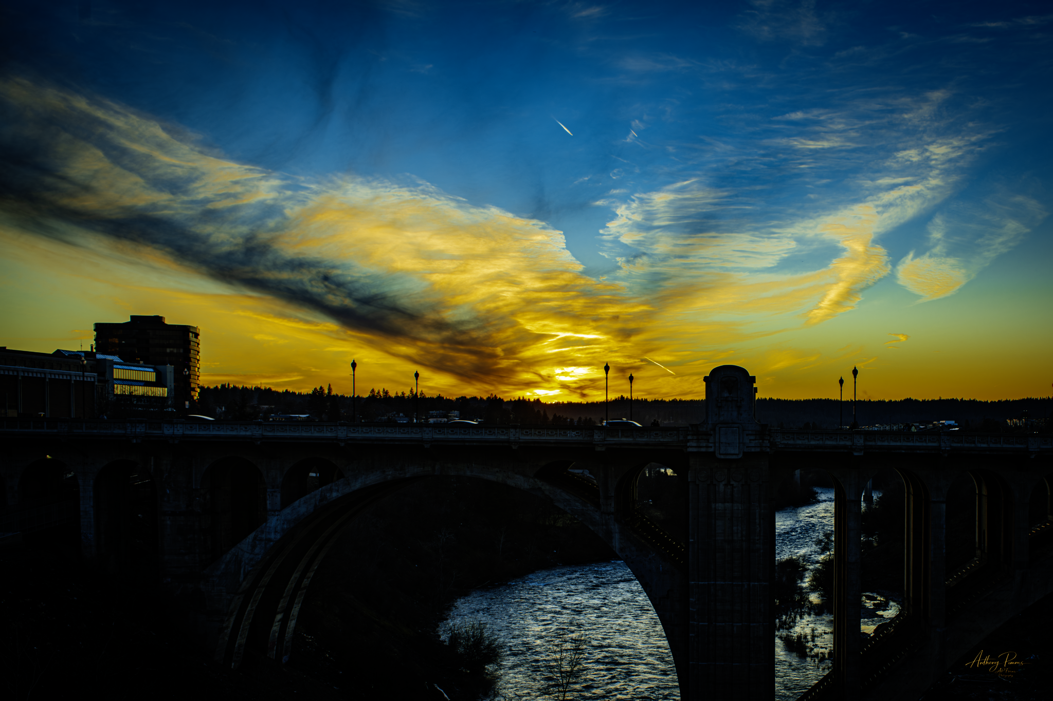 Sunset over a city with a bridge and a river, dramatic clouds in the sky, and a few visible airplanes.